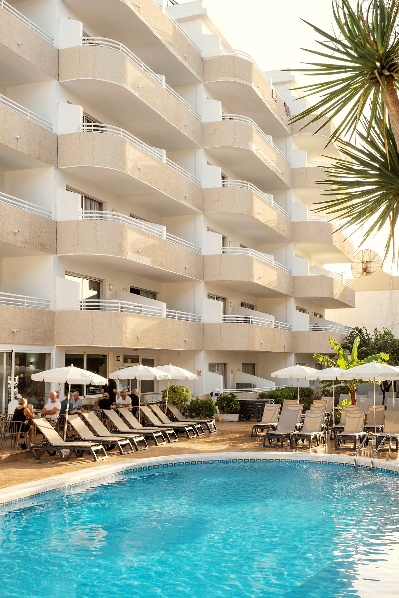 Hotellets facade med altaner og en udendørs pool foran. Liggestole og parasoller står på poolområdet omgivet af grønne områder. (AI-genereret tekst)