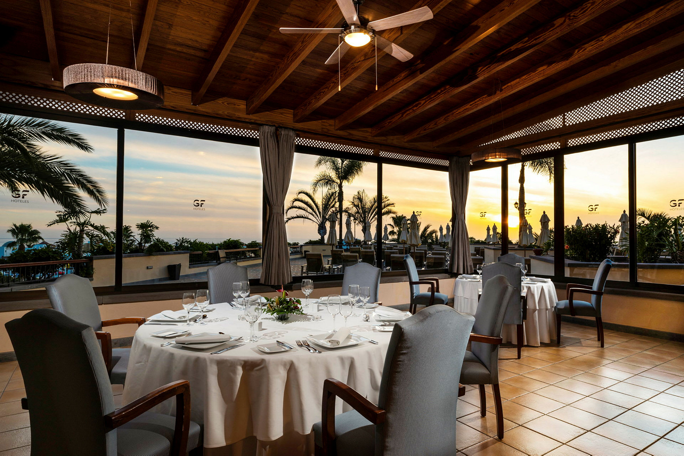Indendørsbillede af en restaurant med dækkede borde og stole, store vinduer med udsigt til udendørsområdet og en solnedgang. (AI-genereret tekst)