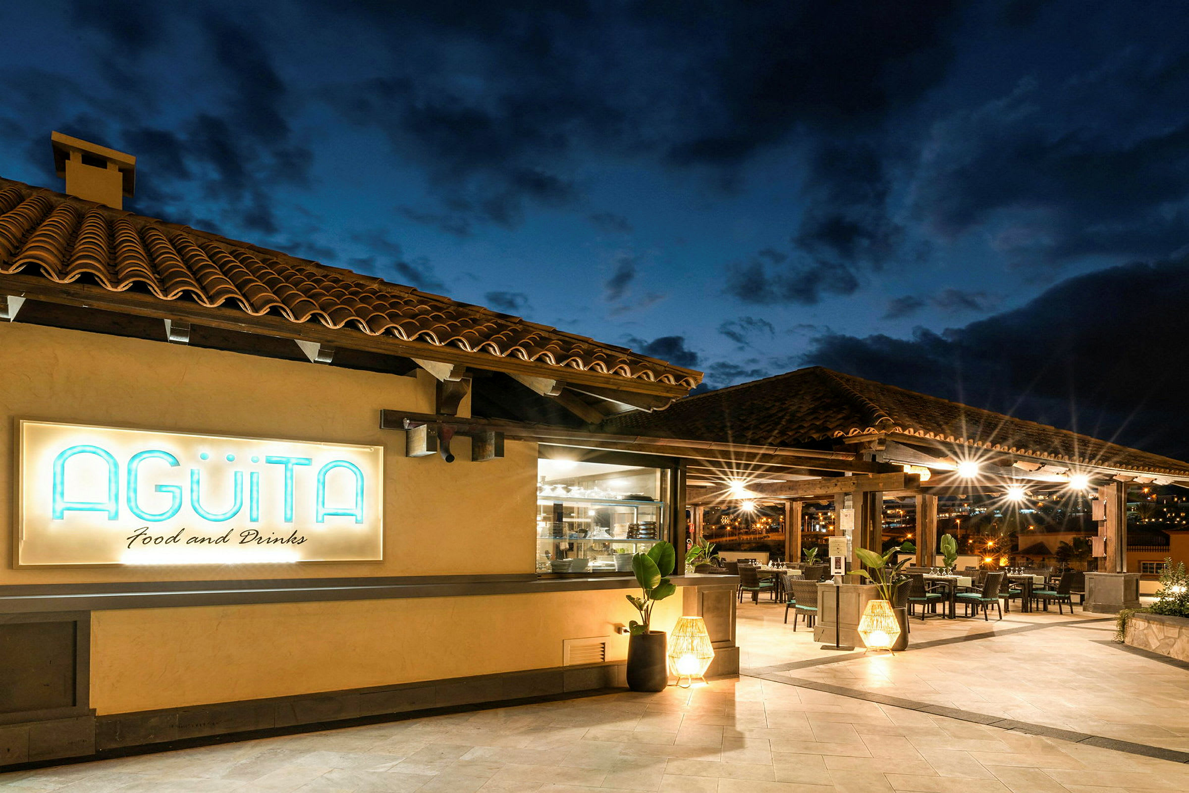 Udendørsbillede af en restaurant med teglstenstag og navnet Agüita belyst på et skilt. (AI-genereret tekst)