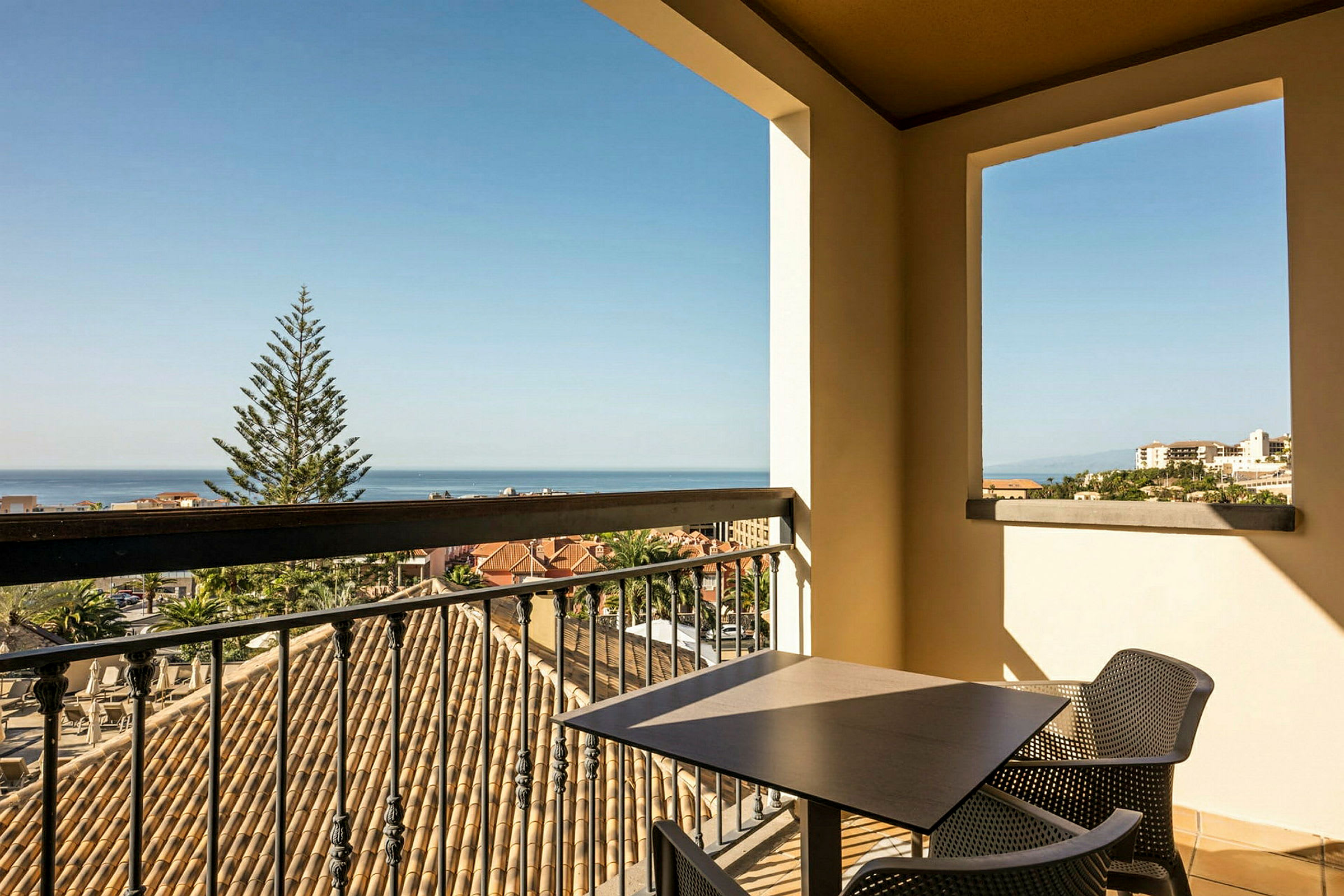 Udsigt fra en balkon med bord og stole over et taglandskab, et træ og havet i baggrunden. (AI-genereret tekst)