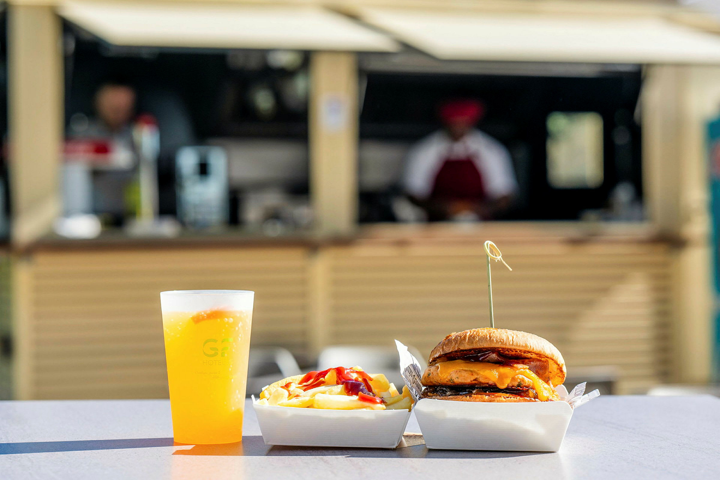Mad bestående af en burger, pommes frites og en drik i en plastkop på et bord udenfor, med en baggrund af en serveringsbod. (AI-genereret tekst)