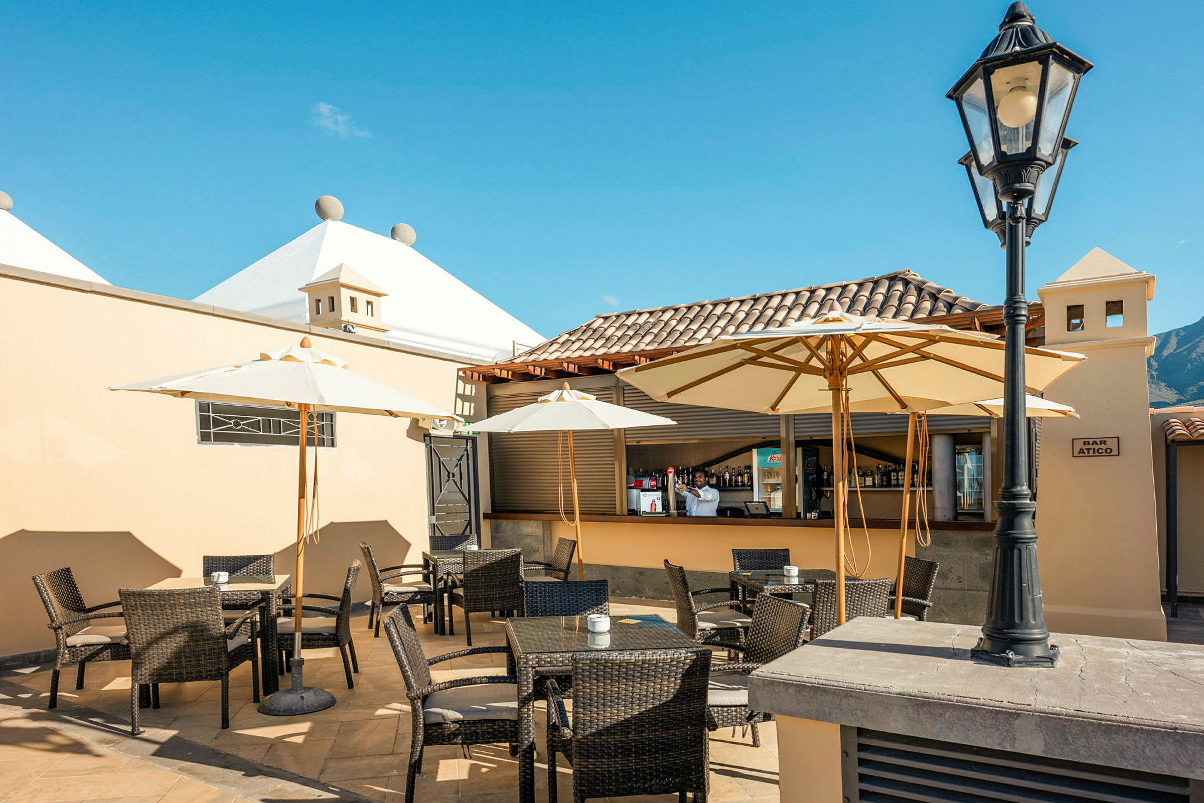 En udendørsrestaurant med borde, stole og parasoller under en klar blå himmel. (AI-genereret tekst)