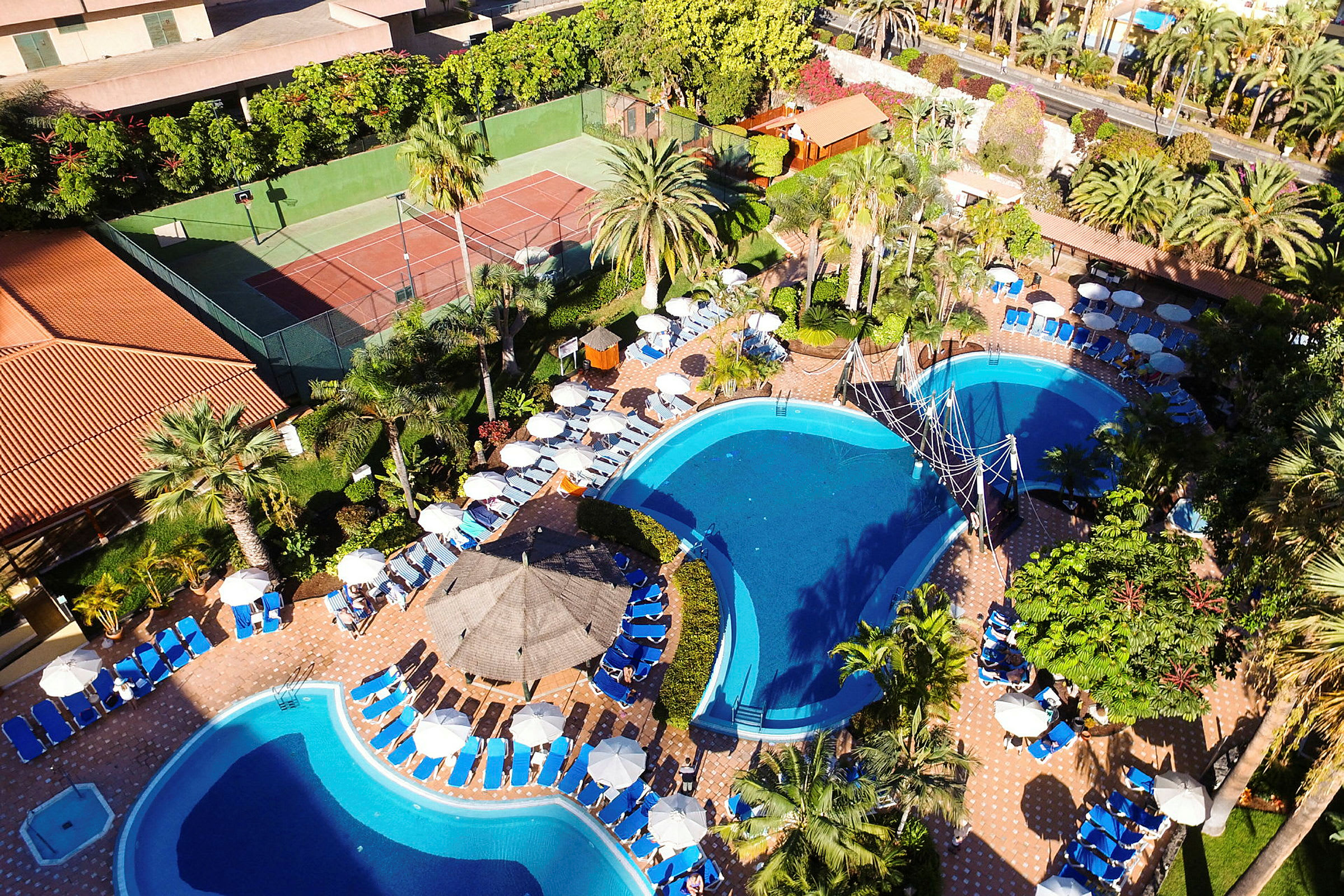 Udsigt over et hotelområde med swimmingpools, liggestole og parasoller i et frodigt område. (AI-genereret tekst)