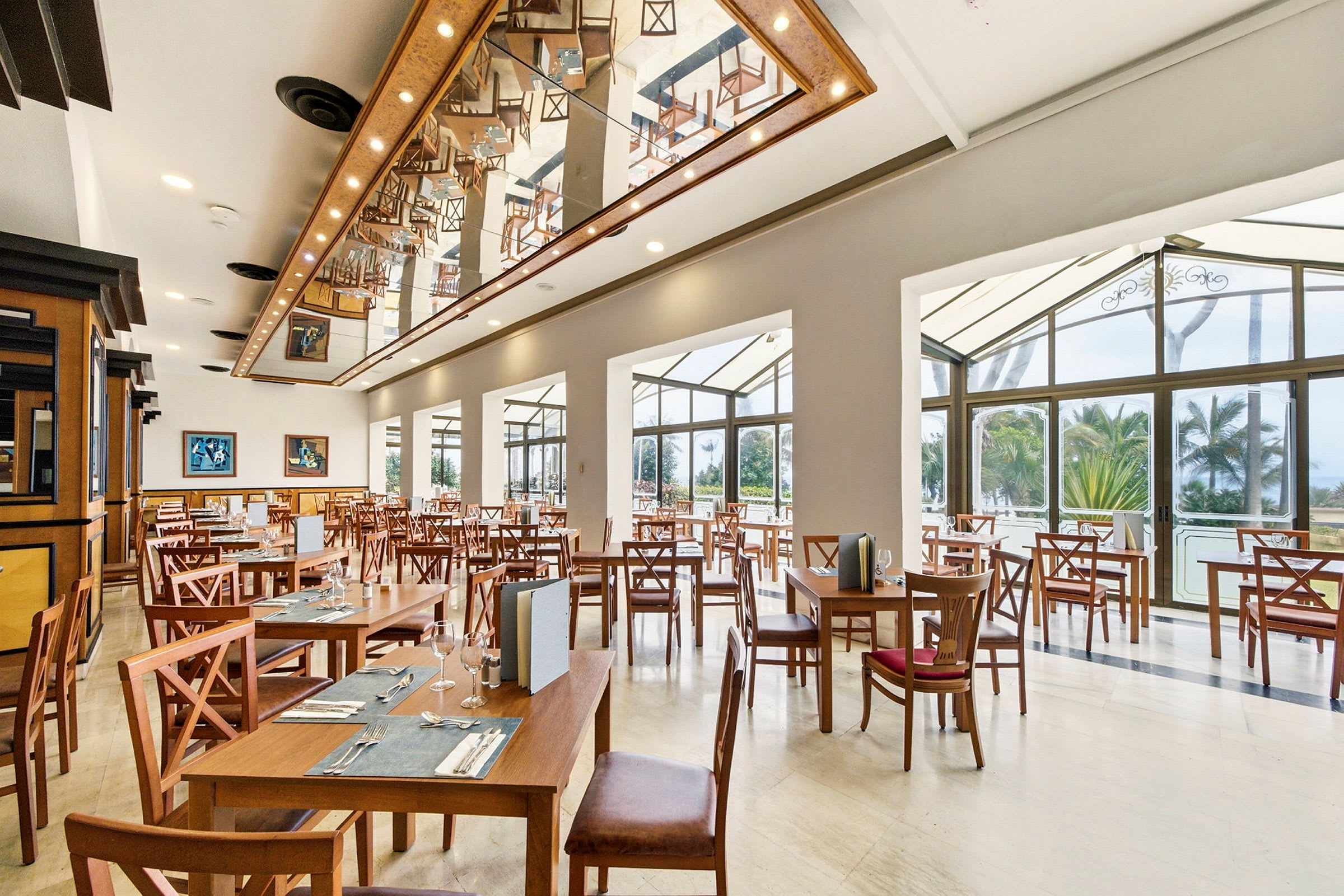 En lys og rummelig restaurant med træborde og -stole, loftsudsmykning og store vinduer med udsigt til grønne omgivelser udenfor. (AI-genereret tekst)