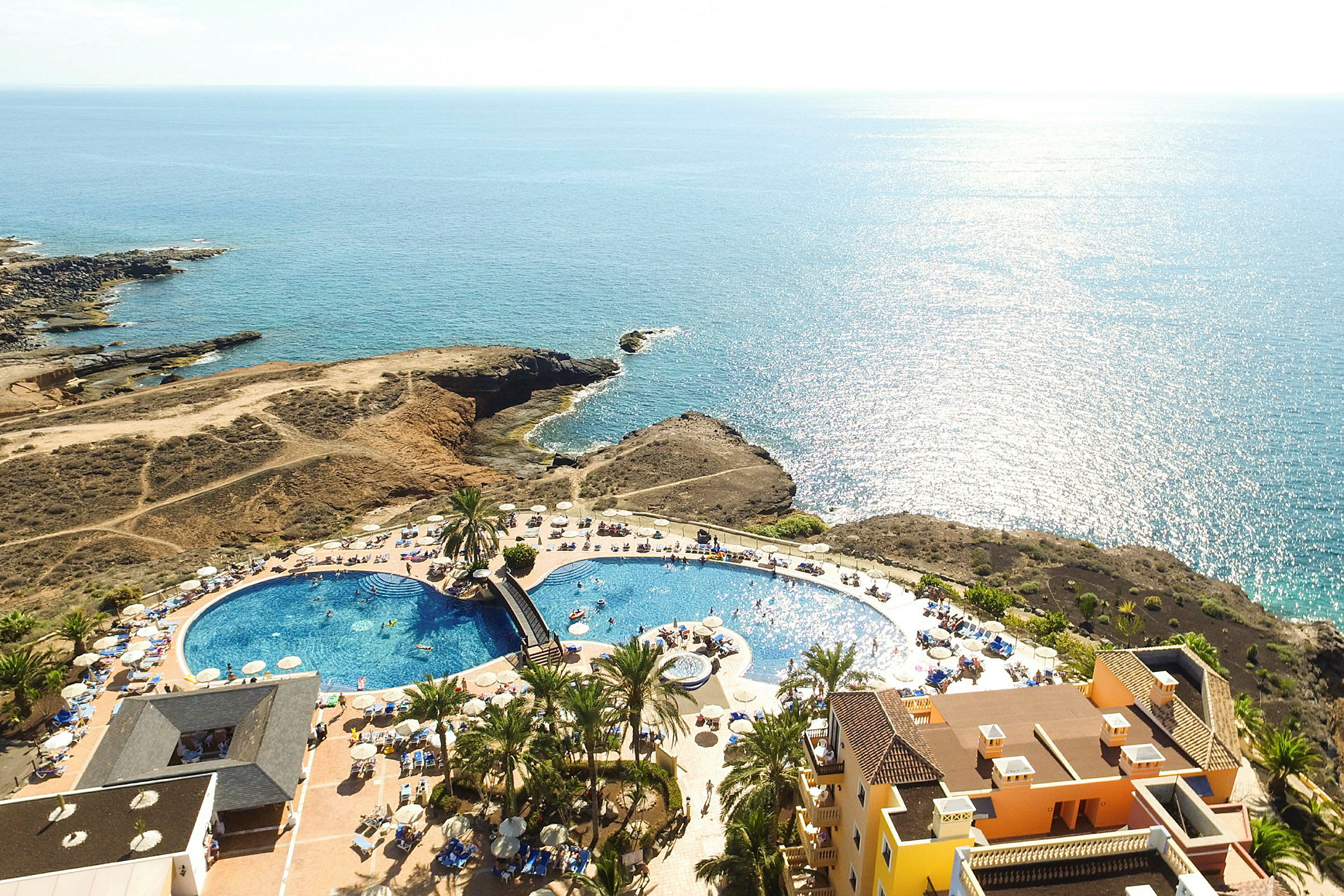 Luftfoto af et resort med swimmingpool, liggestole og havudsigt. (AI-genereret tekst)