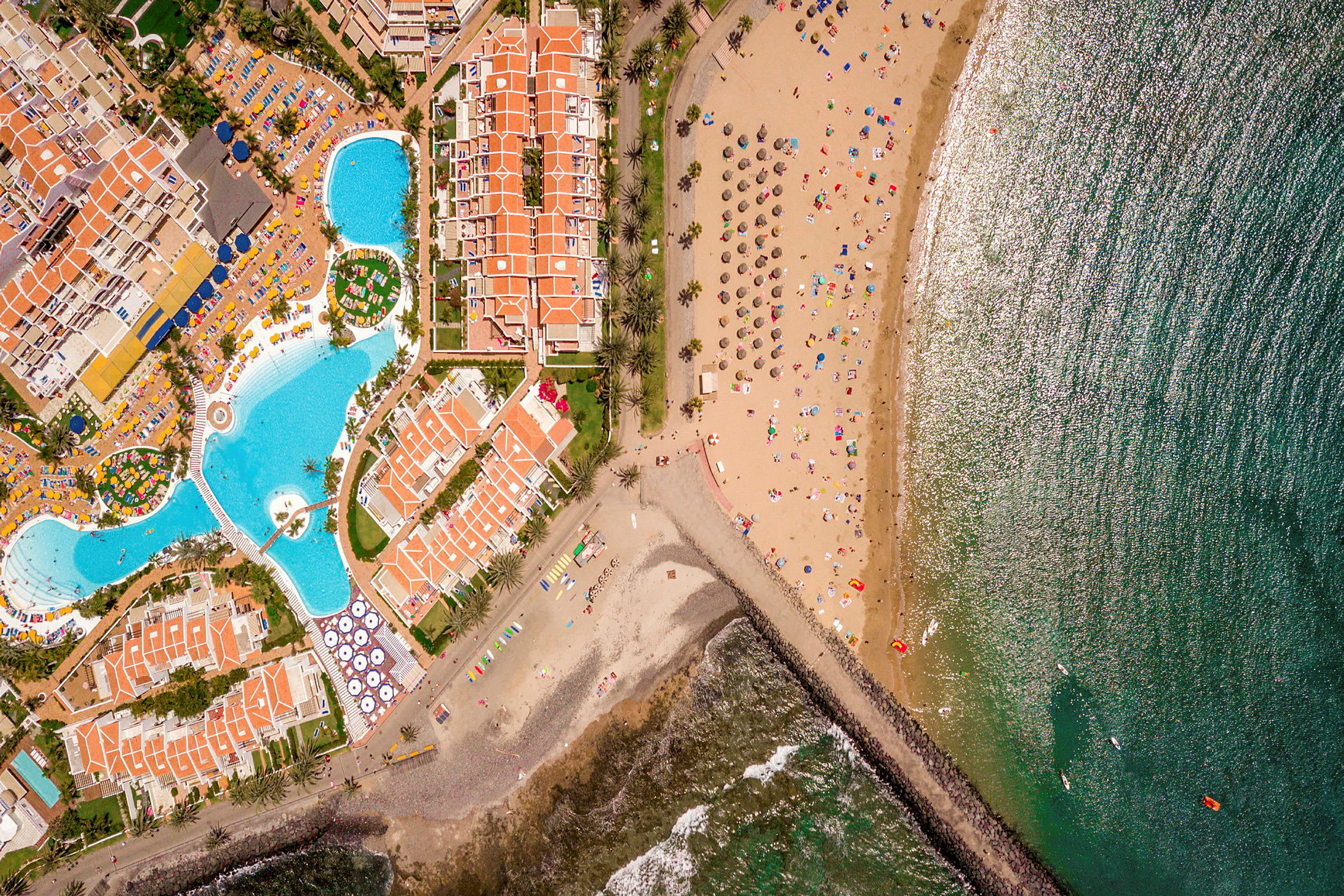 Luftfoto af en strand med solsikringsmidler, en havbugt og tilstødende resorts med swimmingpools. (AI-genereret tekst)