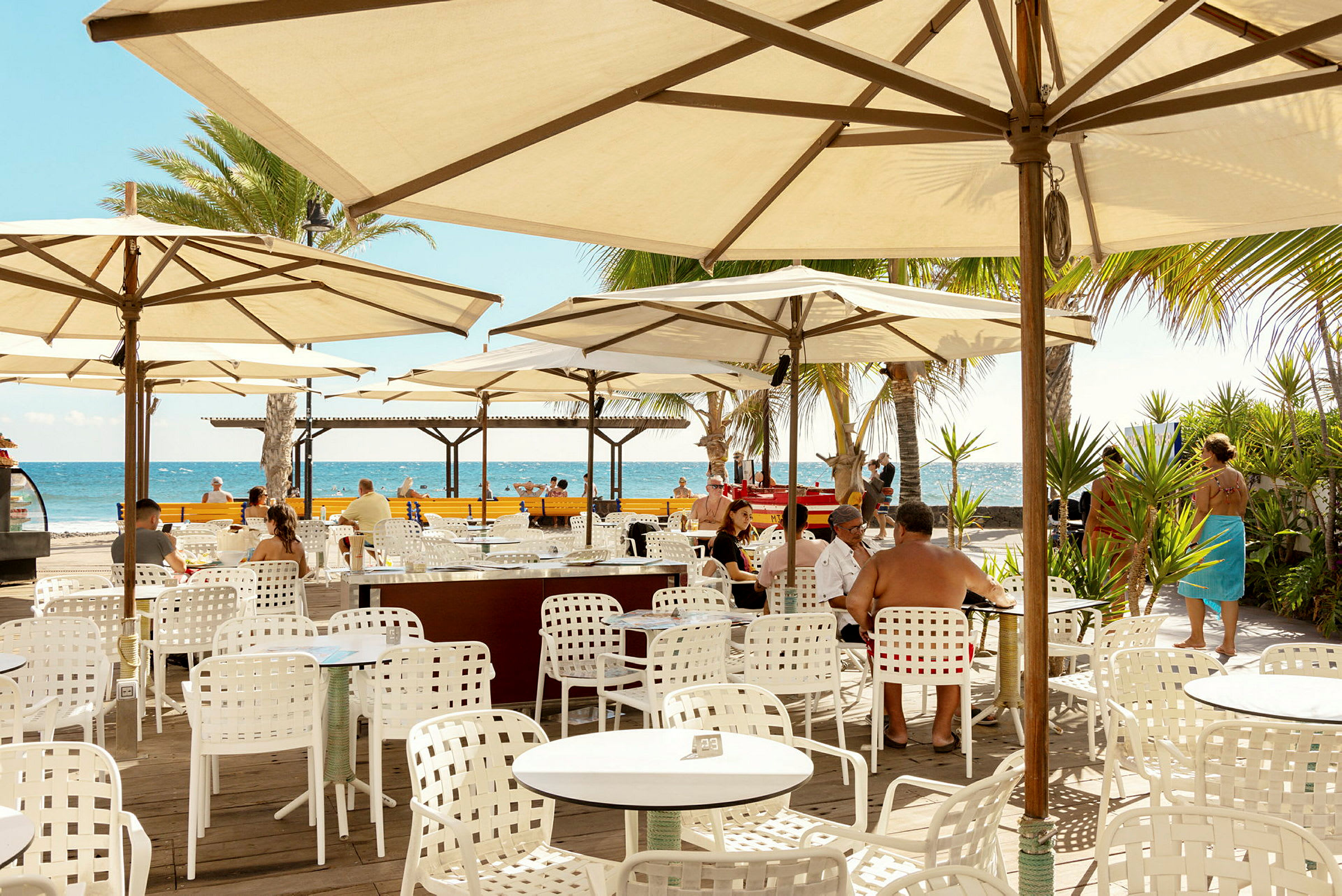 Udendørs restaurant med hvide borde og stole under parasoller, udsigt til havet. (AI-genereret tekst)