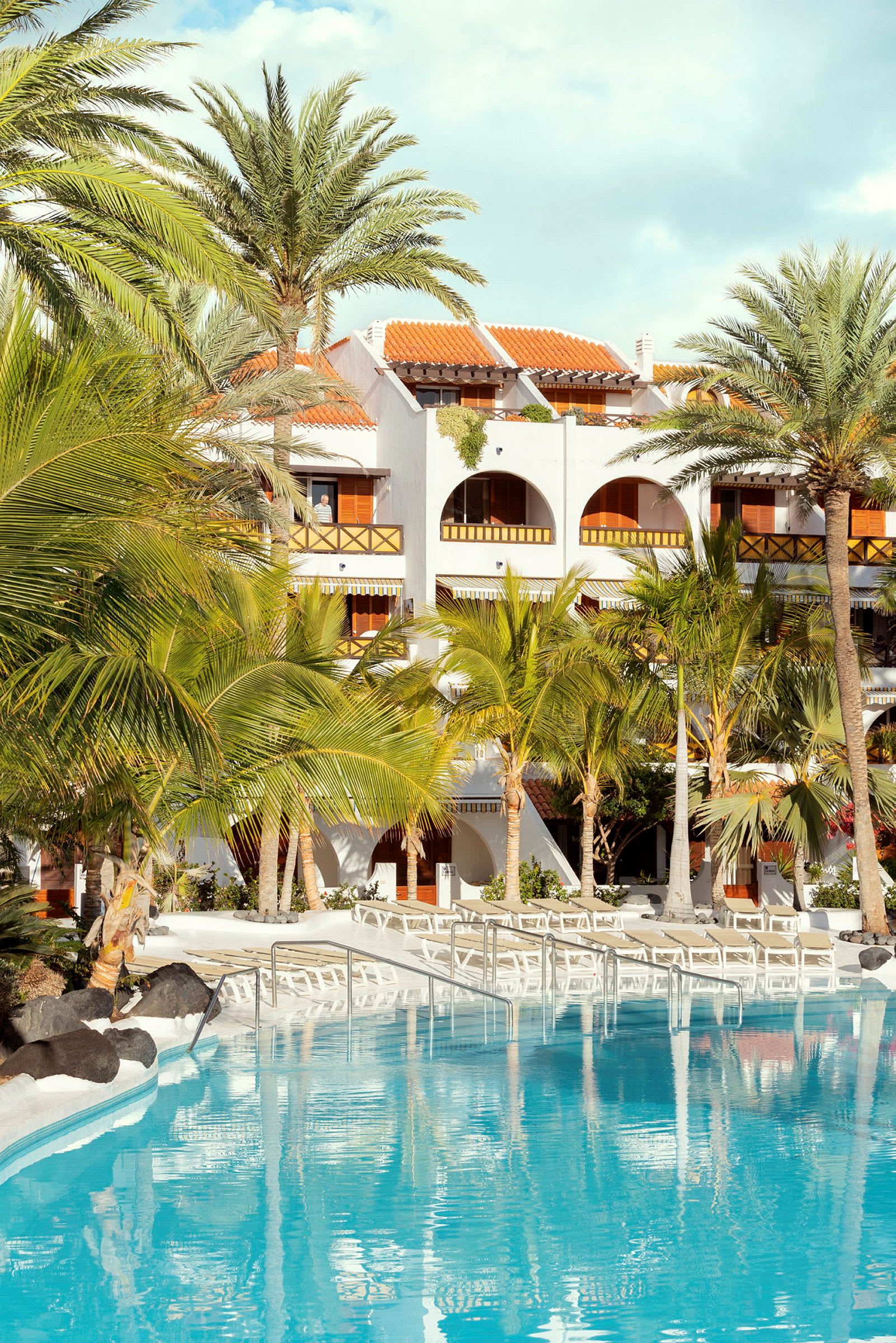 En hotelfacade med flere altaner, omgivet af palmer, med en klar blå pool i forgrunden. (AI-genereret tekst)