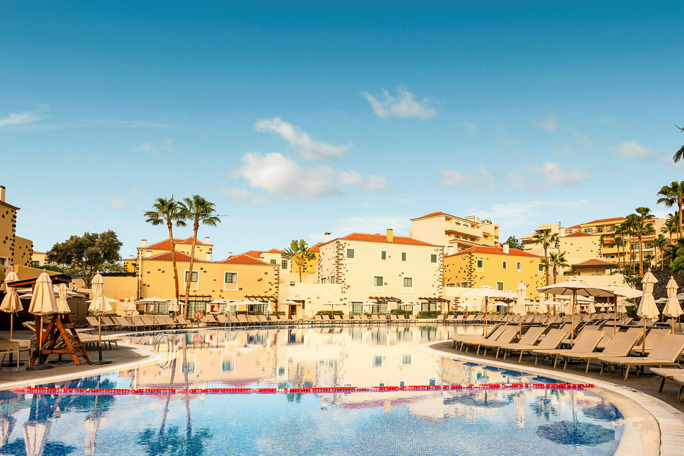 Hotelbygninger med gul facade og røde tage samt en stor swimmingpool omgivet af liggestole og parasoller. (AI-genereret tekst)