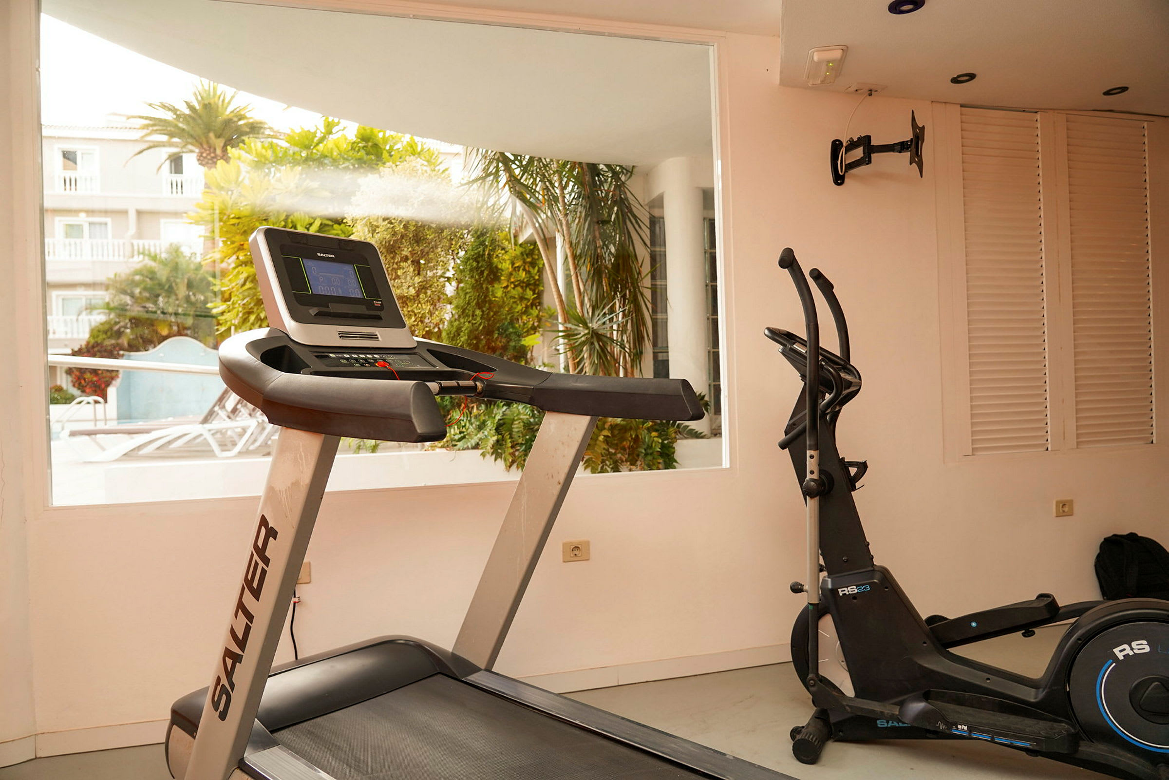 Et fitnessrum med træningsudstyr, herunder et løbebånd og en motionscykel, med udsigt til en have udenfor. (AI-genereret tekst)