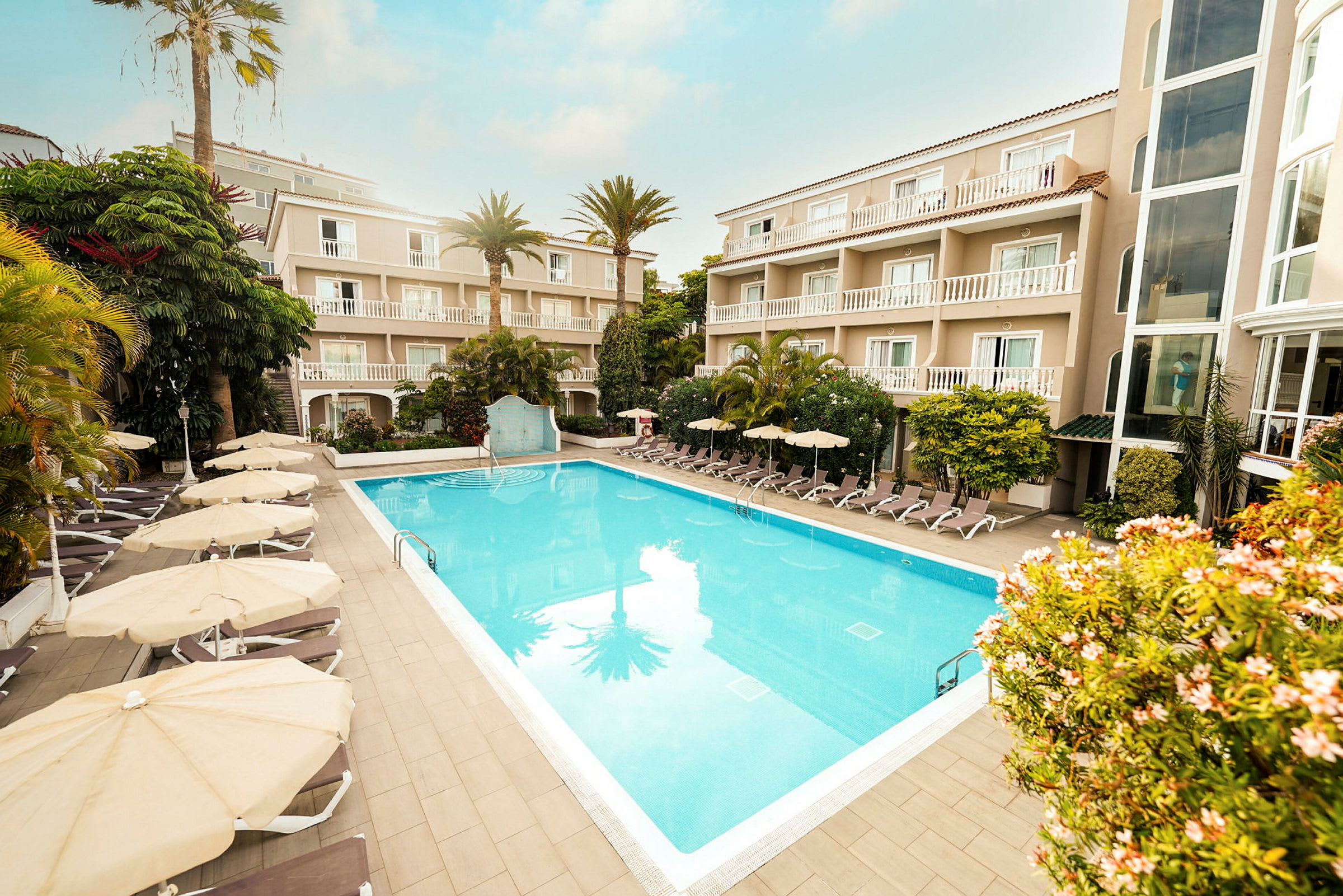 Hotelområde med en udendørs pool omgivet af liggestole og parasoller samt bygninger med altaner. (AI-genereret tekst)