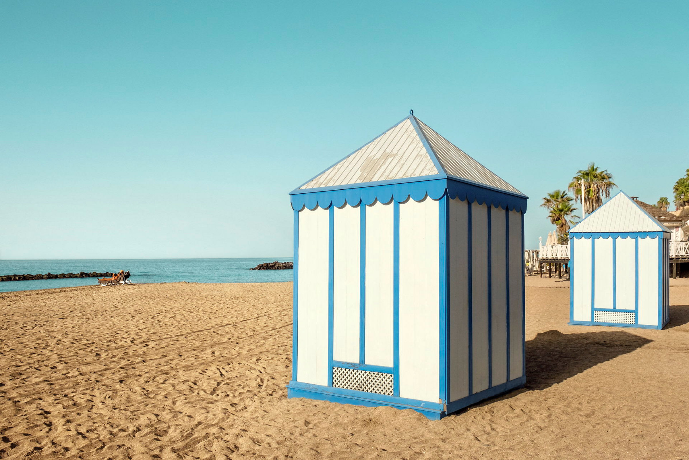 To strandpavilloner med blå og hvide vægge står på en sandstrand ved havet. (AI-genereret tekst)