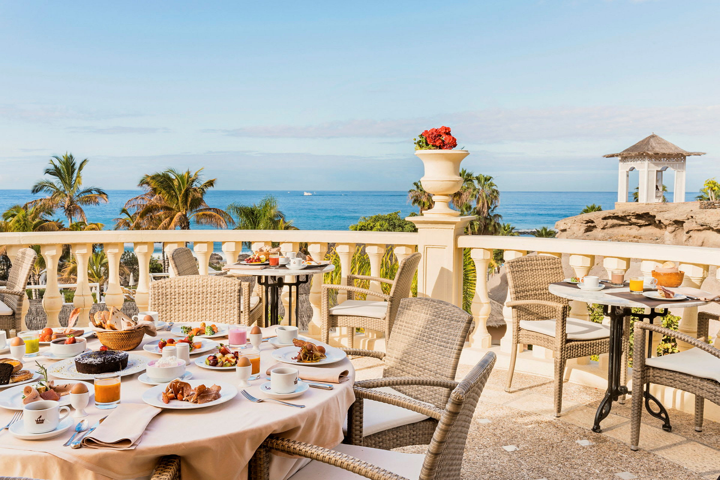 Morgenmadsbord med forskellige retter på en terrasse med udsigt over palmer og havet. (AI-genereret tekst)