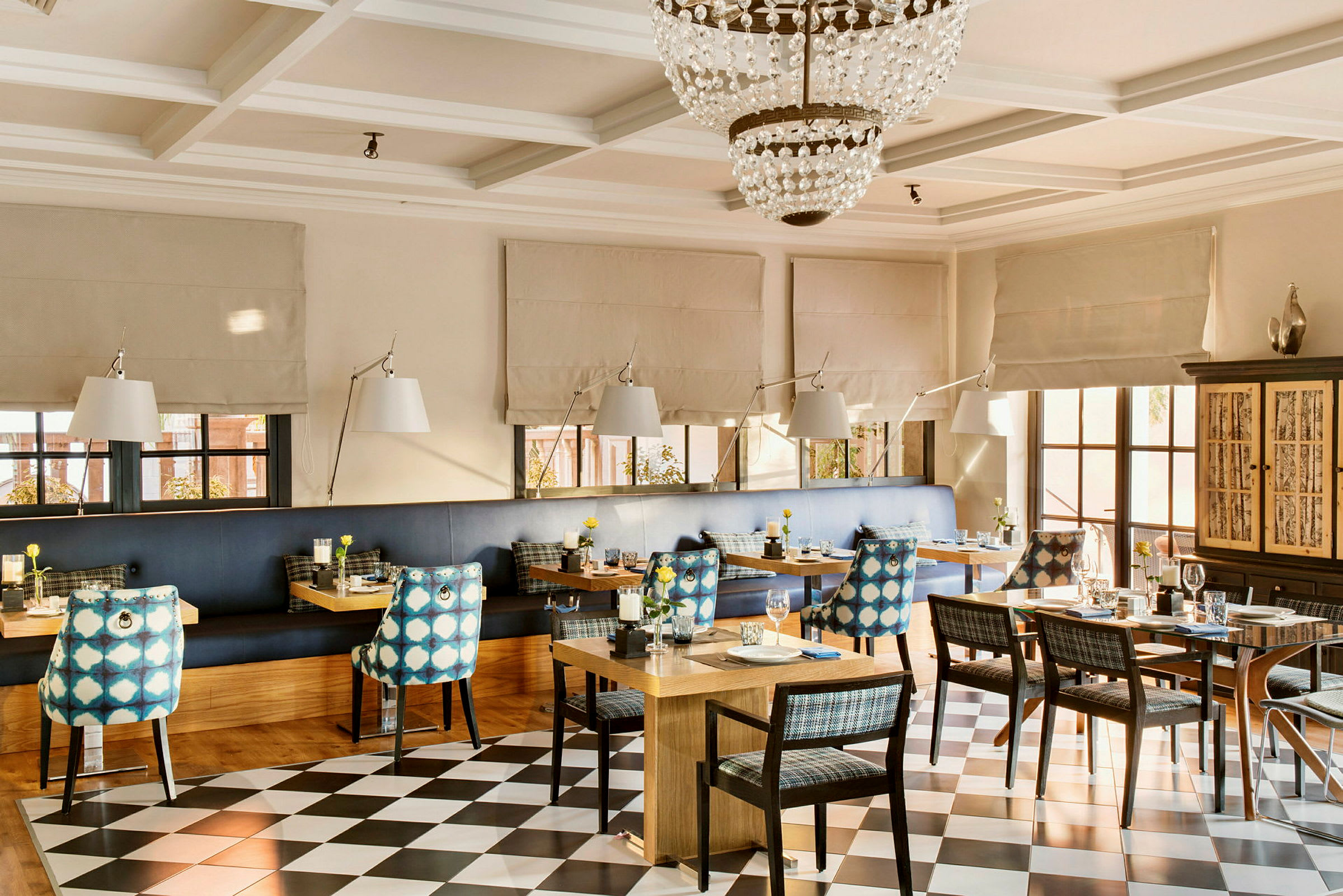 En indendørs restaurant med dækkede borde, stole med mønstrede ryglæn og en lysekrone i loftet. (AI-genereret tekst)