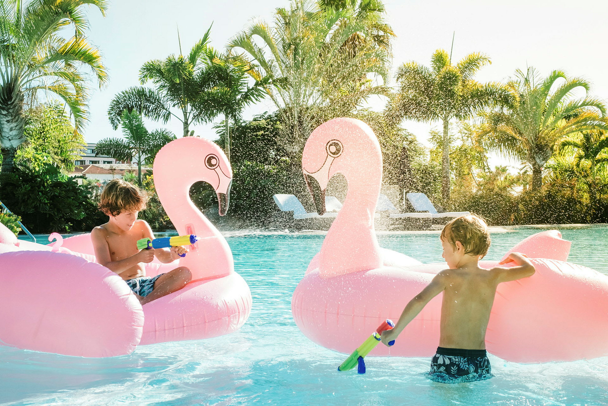 To oppustelige flamingo-figurer i en pool med vandstråler og omgivelser med palmer. (AI-genereret tekst)