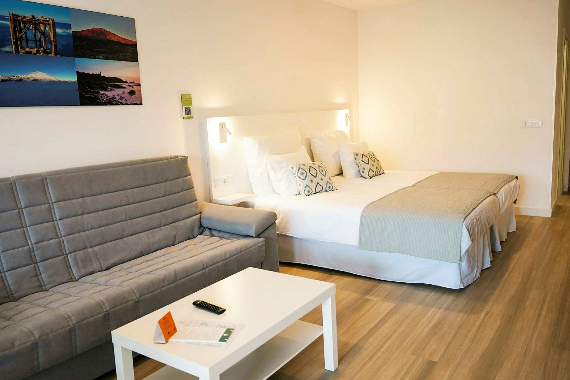 Hotelværelse med en dobbeltseng, en grå sofa, et hvidt bord og billeder på væggen. (AI-genereret tekst)