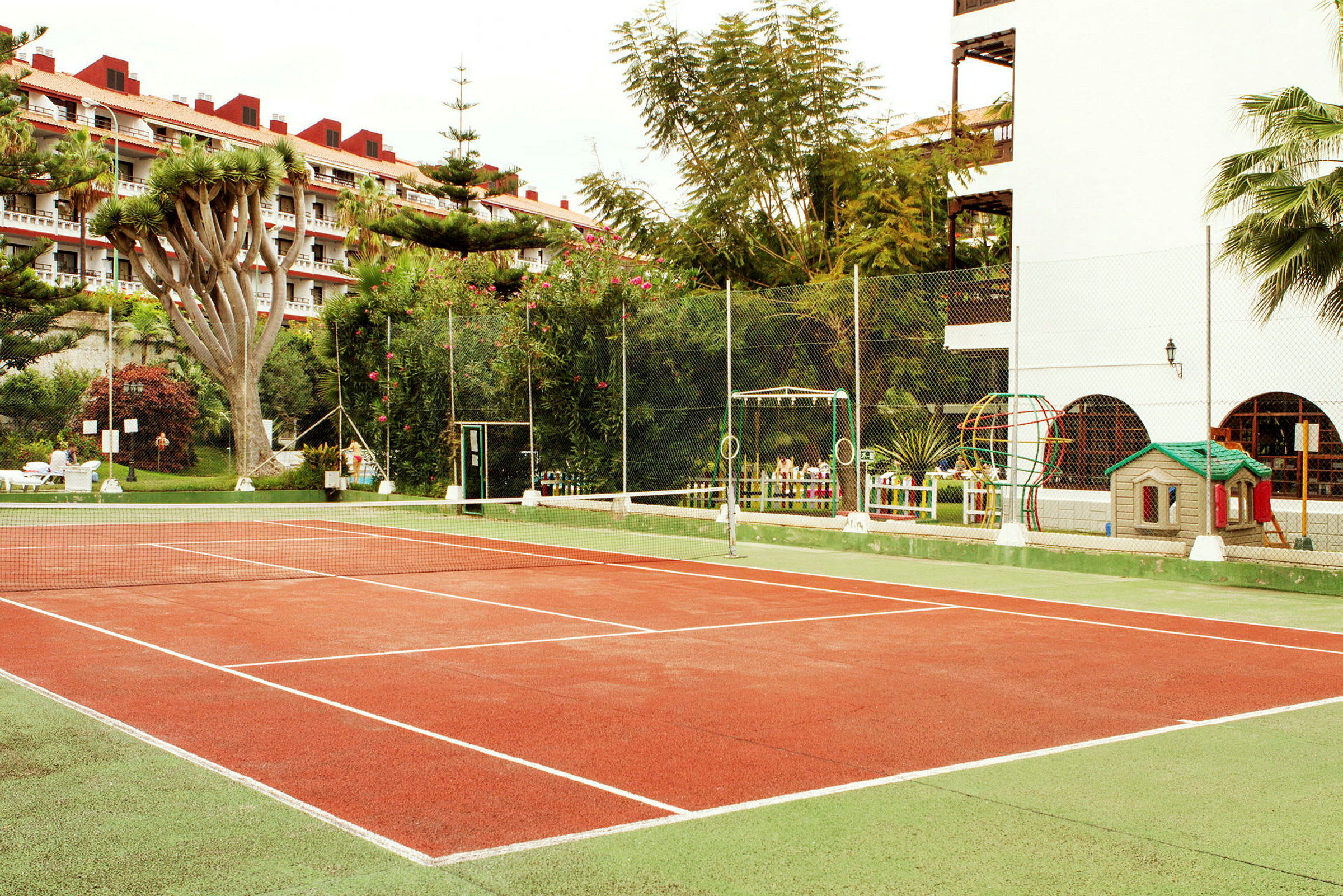 En tennisbane omgivet af beplantning og bygninger. (AI-genereret tekst)