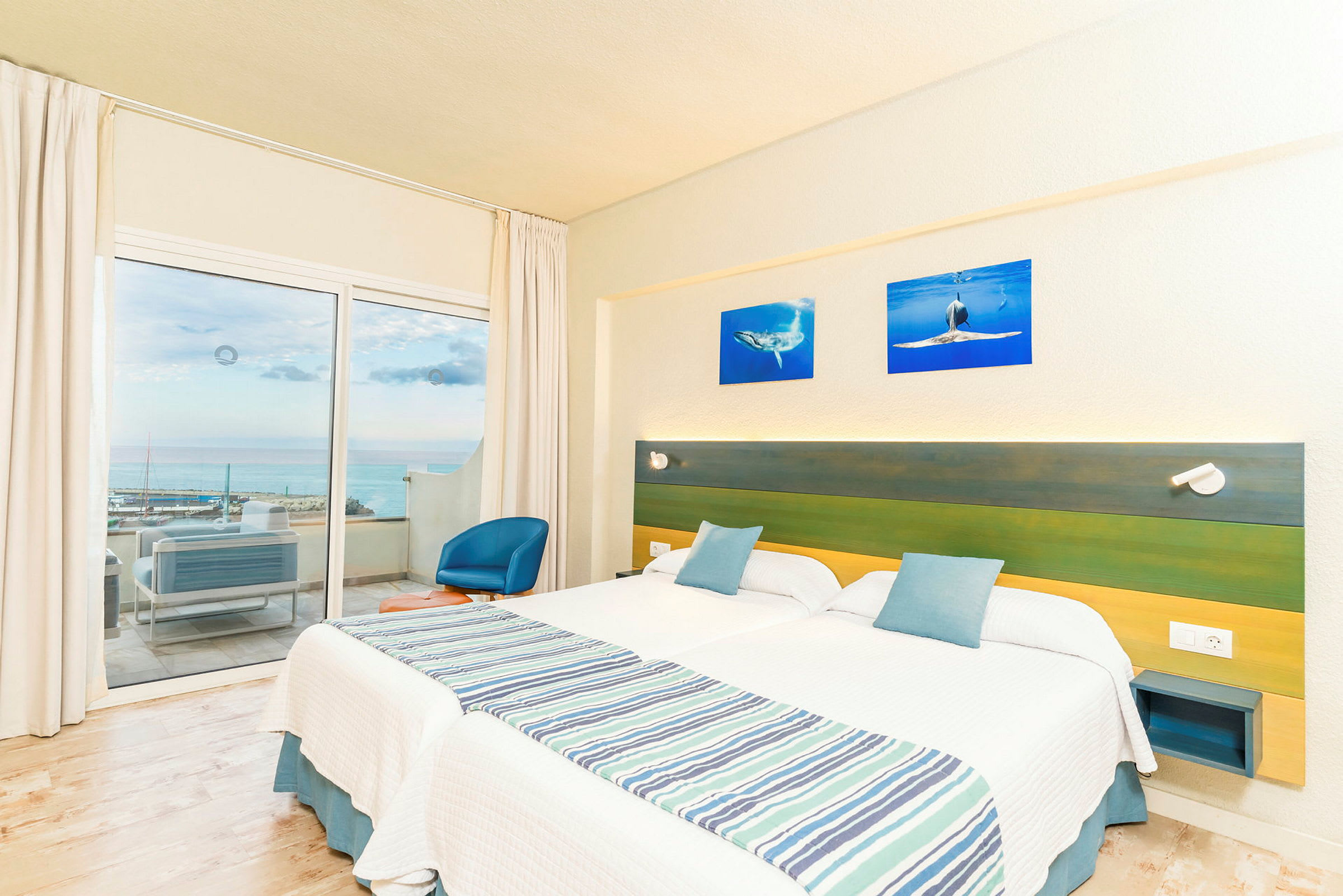 Hotelværelse med to enkeltsenge, en grøn og blå sengegavl samt en balkon med udsigt over havet. (AI-genereret tekst)