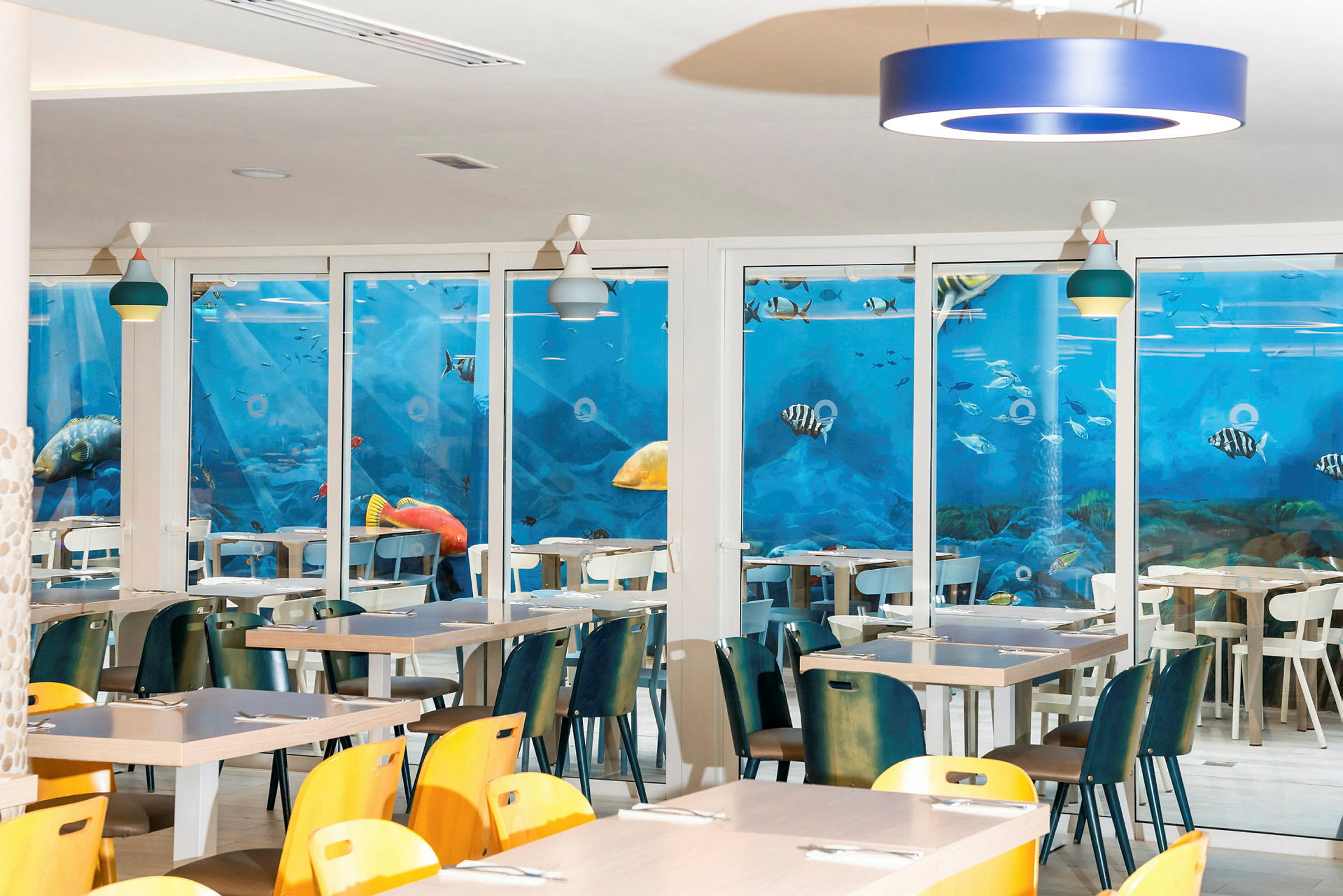 Restaurant med borde og stole, med en væg dækket af et blåt akvariemotiv. (AI-genereret tekst)