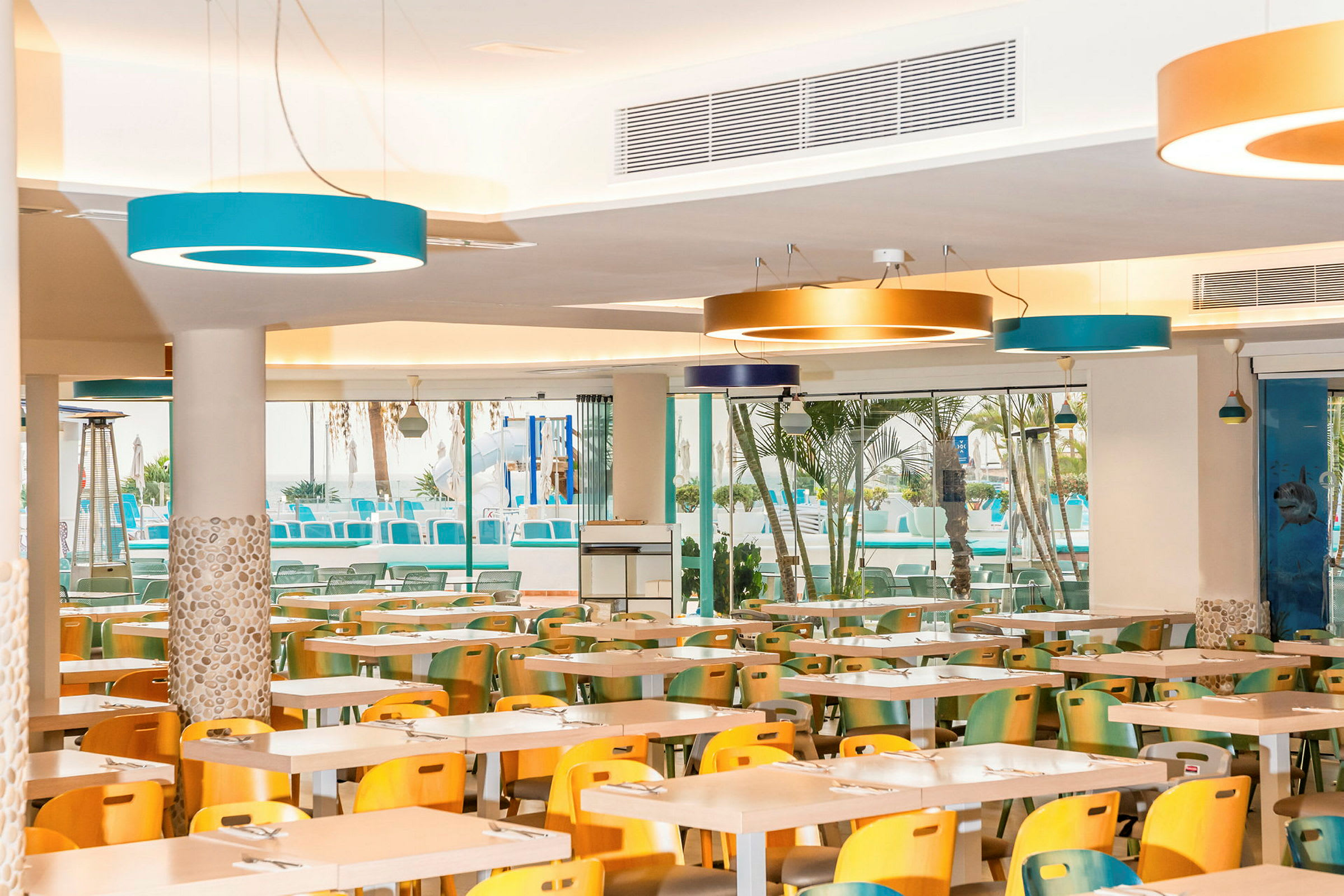 En restaurant med farverige stole og borde, samt store vinduer mod en udendørs pool. (AI-genereret tekst)