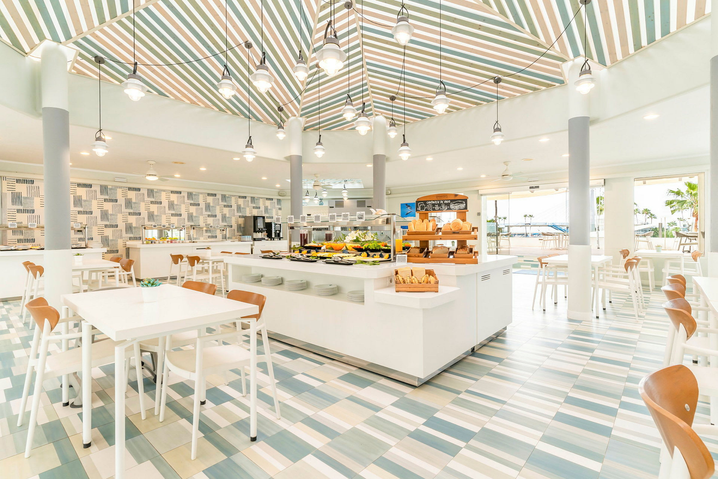 En lys og moderne restaurant med hvide møbler og farverige fliser på gulvet. Et buffetbord er centralt placeret med forskellige retter. (AI-genereret tekst)