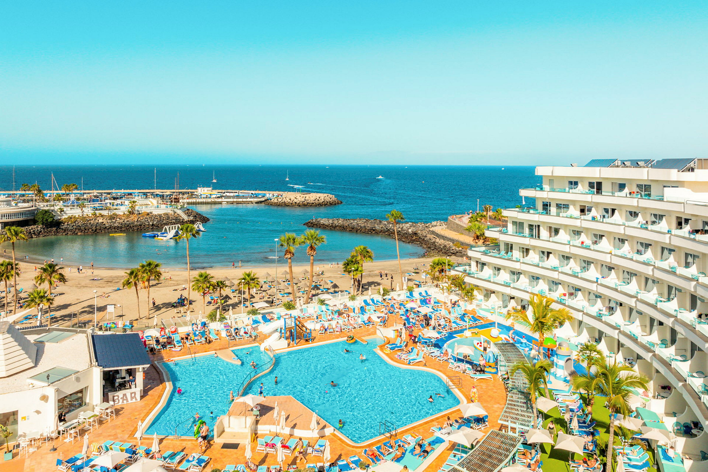 Billede af et hotel med poolområde, strand og havneindløb ved havet. (AI-genereret tekst)