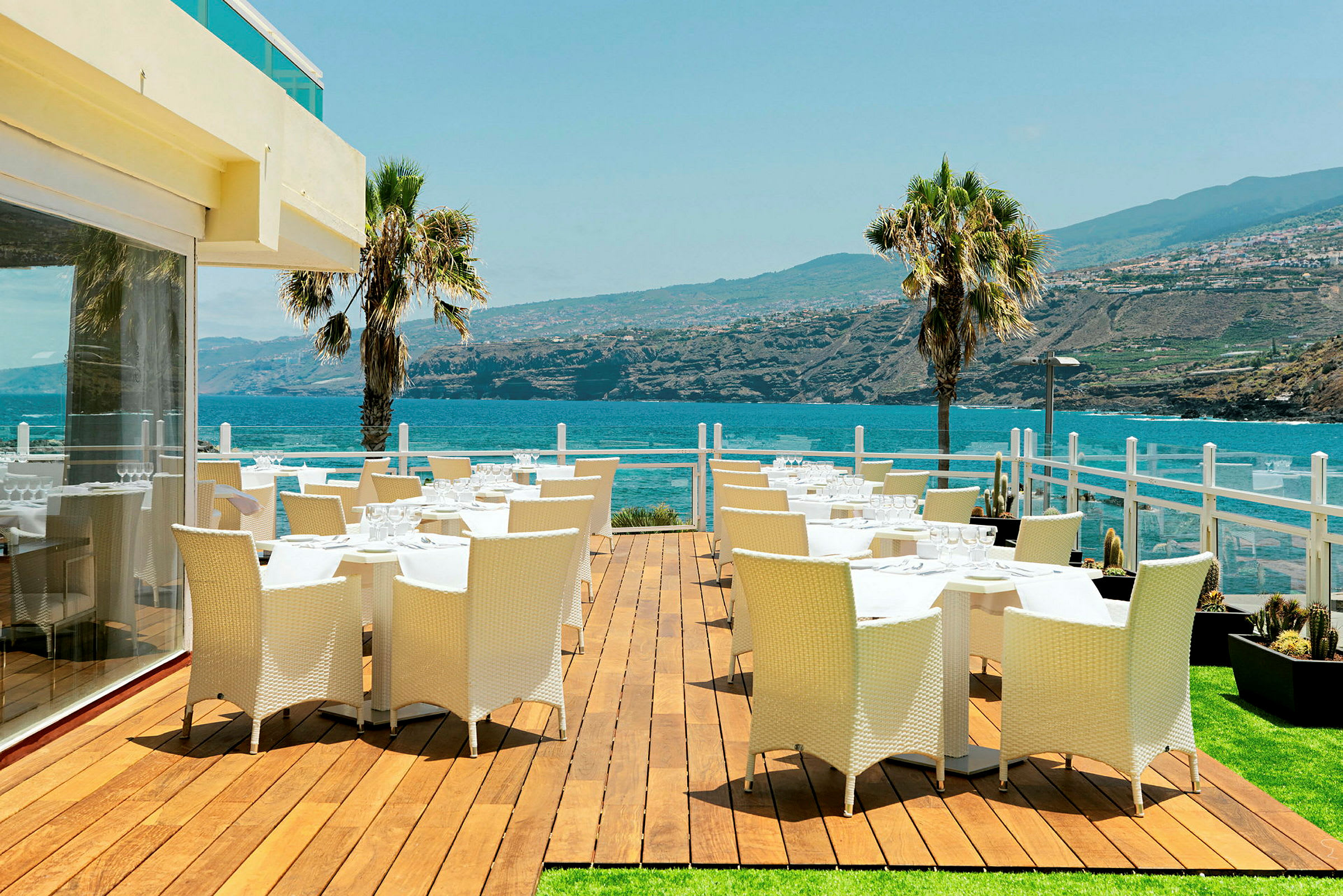 Udendørs terrasse med spiseborde og stole, udsigt over havet og det omkringliggende landskab. (AI-genereret tekst)
