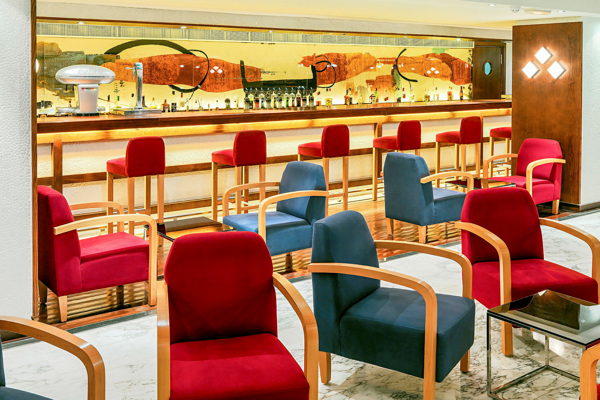 En hotelbar med oplyst bardisk, høje røde stole og komfortable siddepladser i forskellige farver. (AI-genereret tekst)