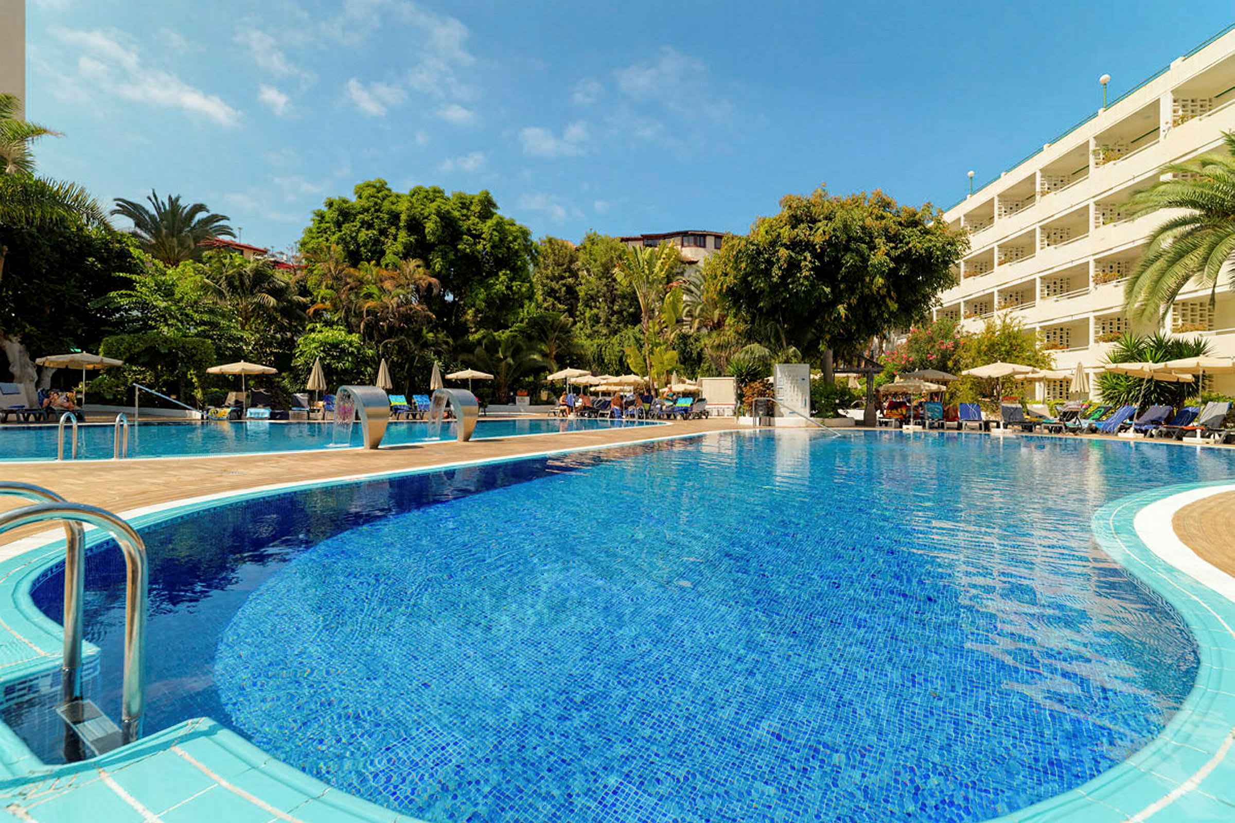 En solrig hotelpool omgivet af grønne områder og liggestole. (AI-genereret tekst)