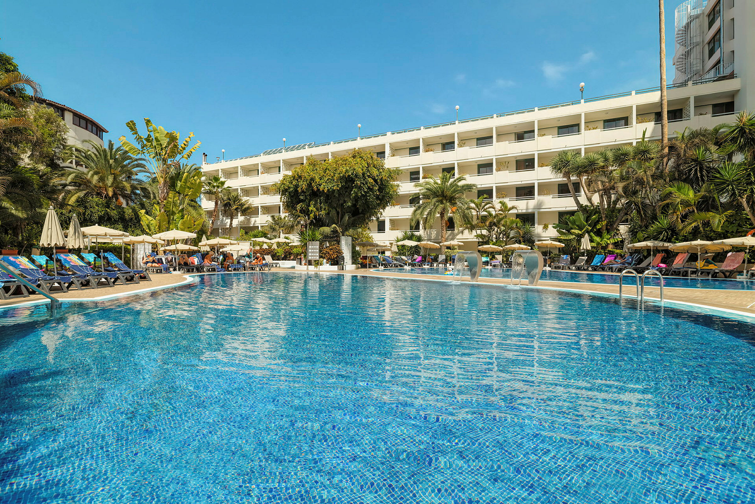 En swimmingpool foran et hotel omgivet af palmer og liggestole. (AI-genereret tekst)