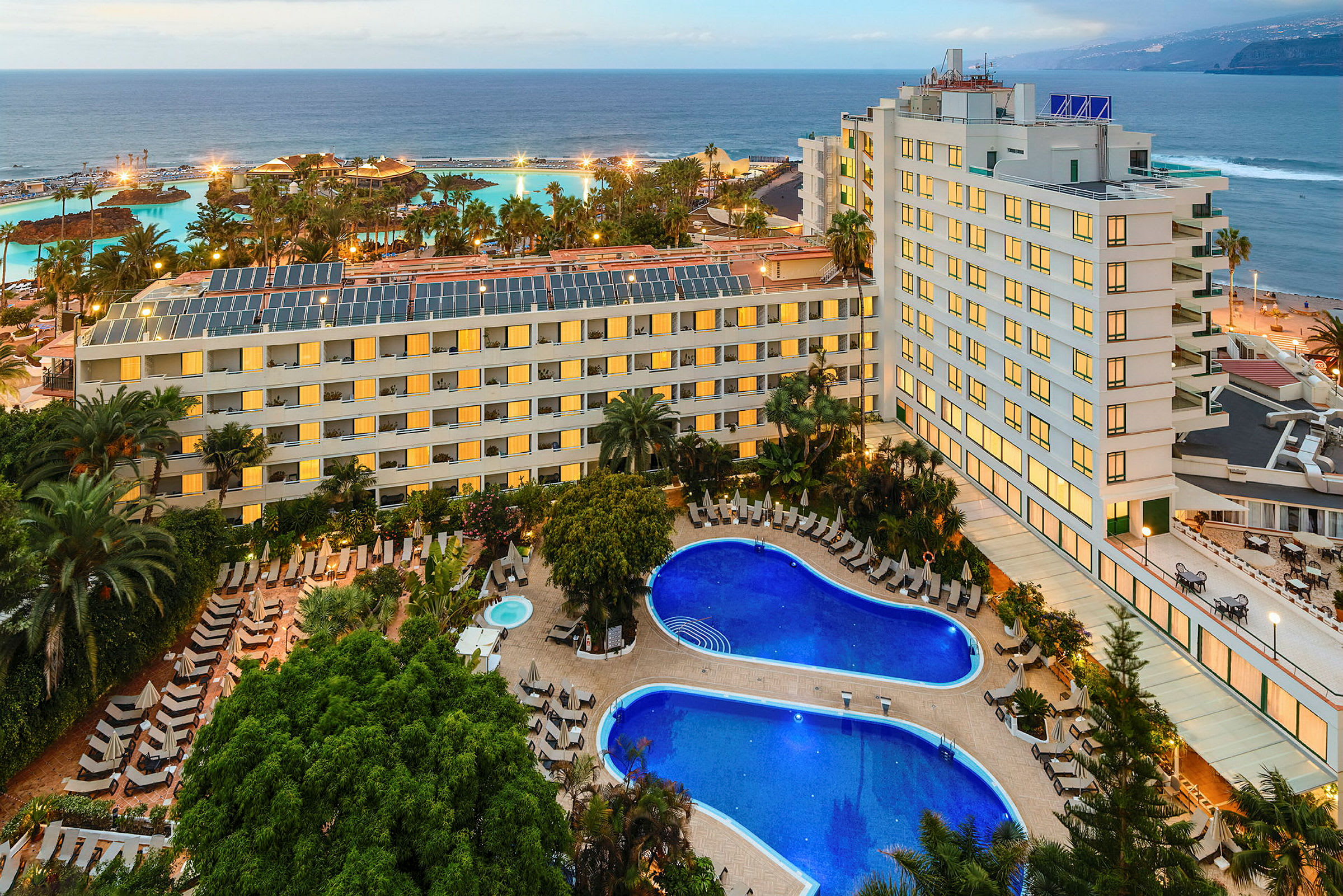 Et hotelkompleks med pools og grønne områder i forgrunden samt en kystlinje og hav i baggrunden. (AI-genereret tekst)