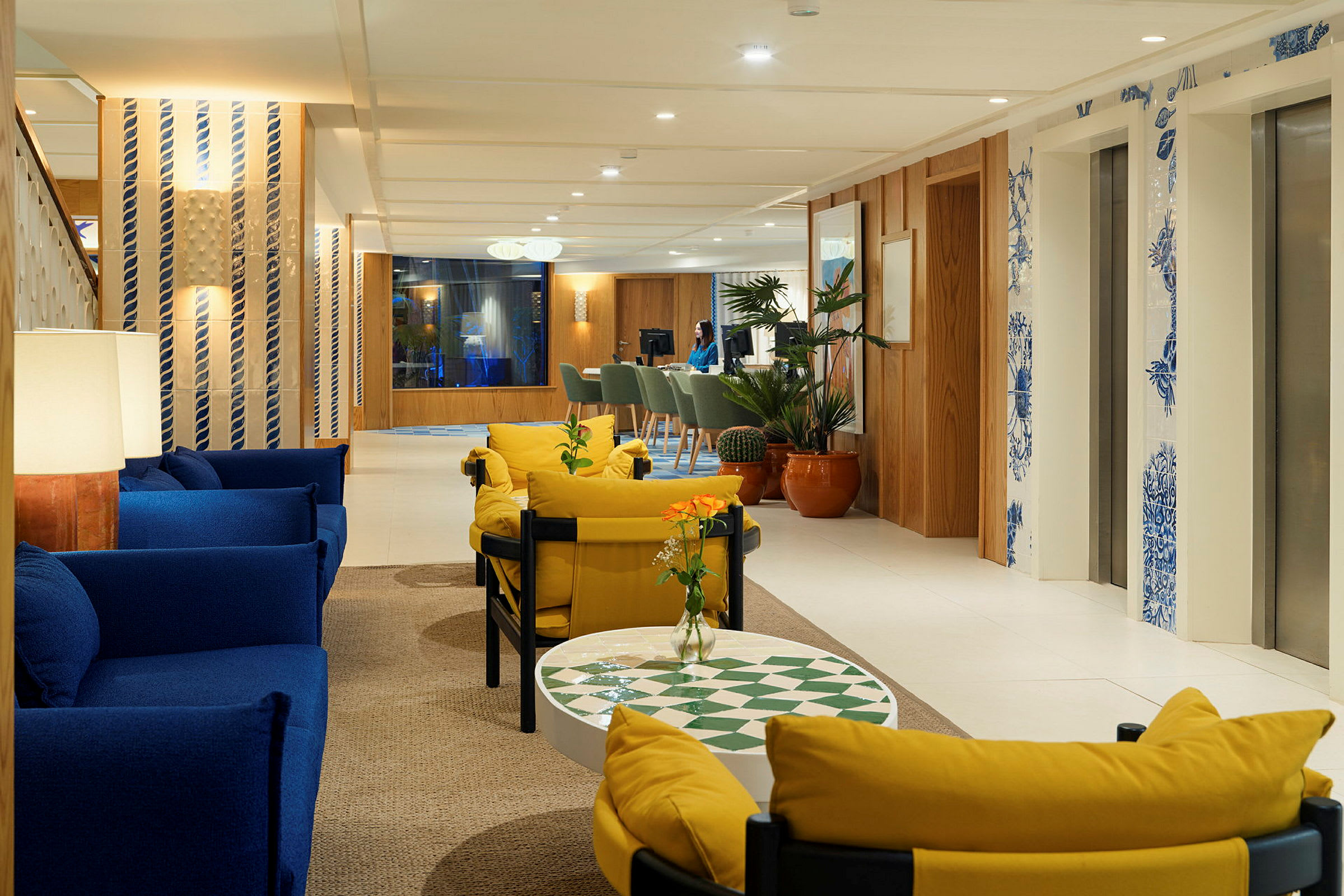 En lys hotellobby med sofaer i blå og gul, planter og elevatorer dekoreret med blå detaljer. (AI-genereret tekst)