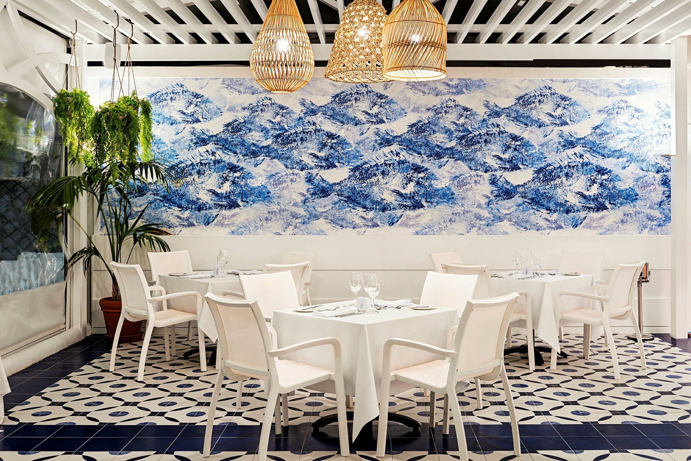 En indendørs restaurant med hvide borde og stole, dekoreret med et stort vægmaleri i blå og hvidt, der forestiller bjerge. Hængende planter pryder lokalet. (AI-genereret tekst)