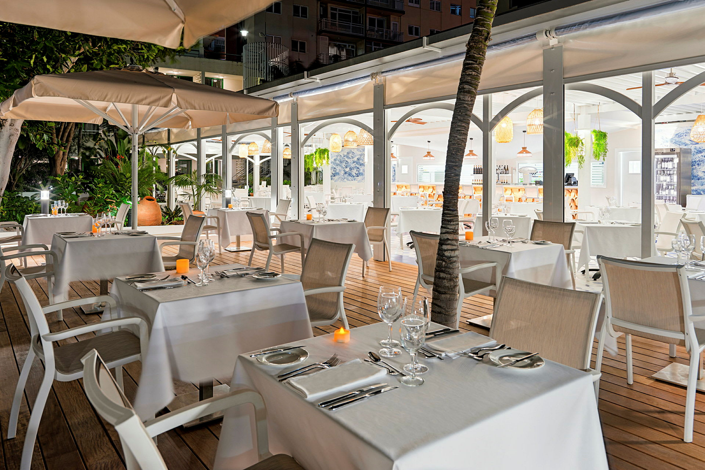 En udendørs restaurant med dækkede borde og stole, omgivet af grønt og oplyst af blødt lys. (AI-genereret tekst)