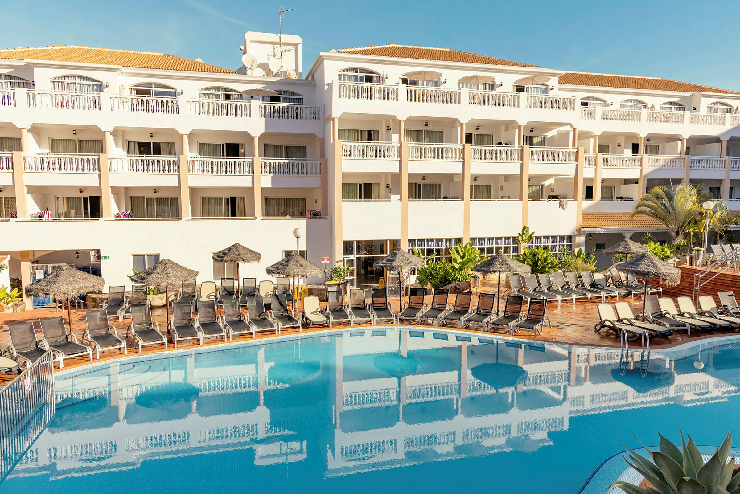 Hotelbygning med altaner og en swimmingpool omgivet af liggestole. (AI-genereret tekst)