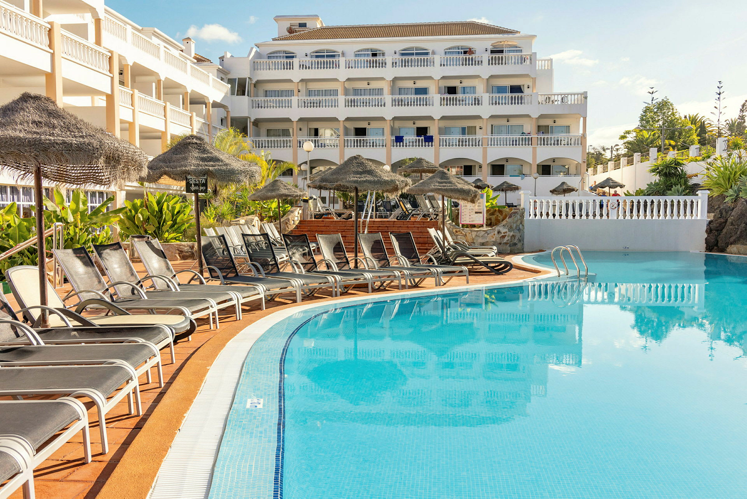 Billede af en hotelbygning med flere etager beliggende ved en pool. Foran poolen står liggestole og parasoller. (AI-genereret tekst)
