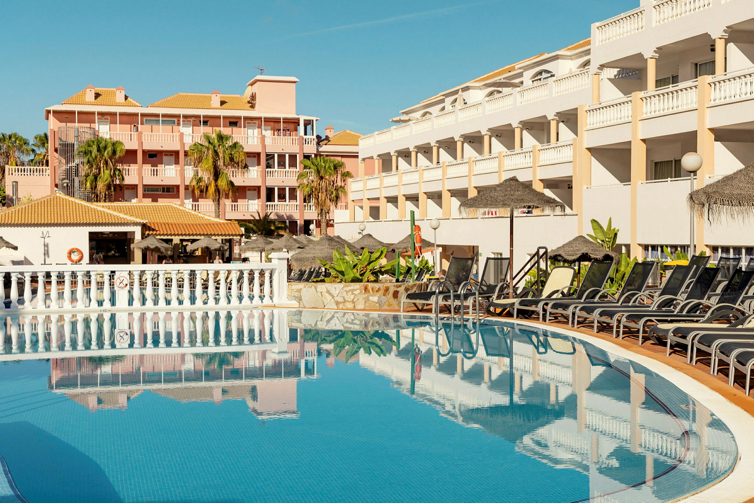 Billede af en udendørs pool med liggestole og parasoller ved et hotel. (AI-genereret tekst)