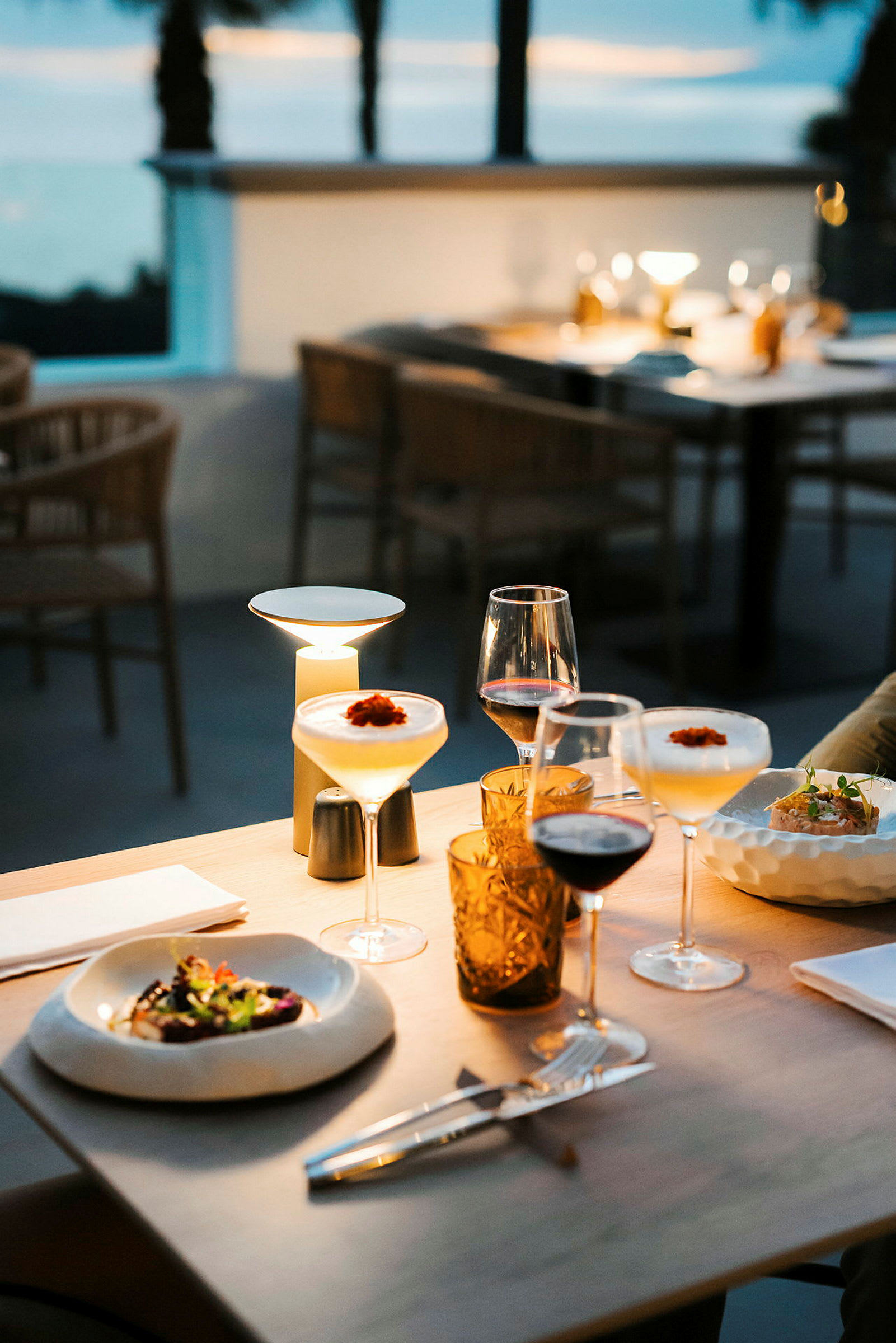 Et bord på en udendørsrestaurant med mad og drikke serveret, omgivet af en stemningsfuld aftenbelysning. (AI-genereret tekst)
