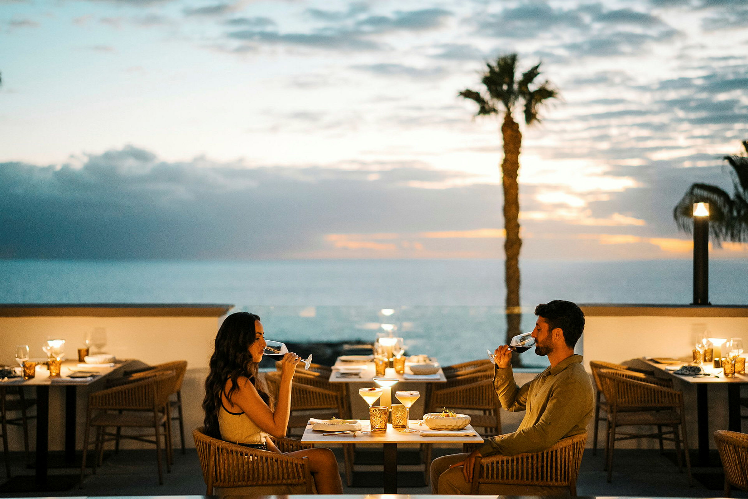 To personer sidder ved et dækket bord på en udendørs restaurant og nyder et glas med drikke. I baggrunden ses havet og en solnedgang. (AI-genereret tekst)