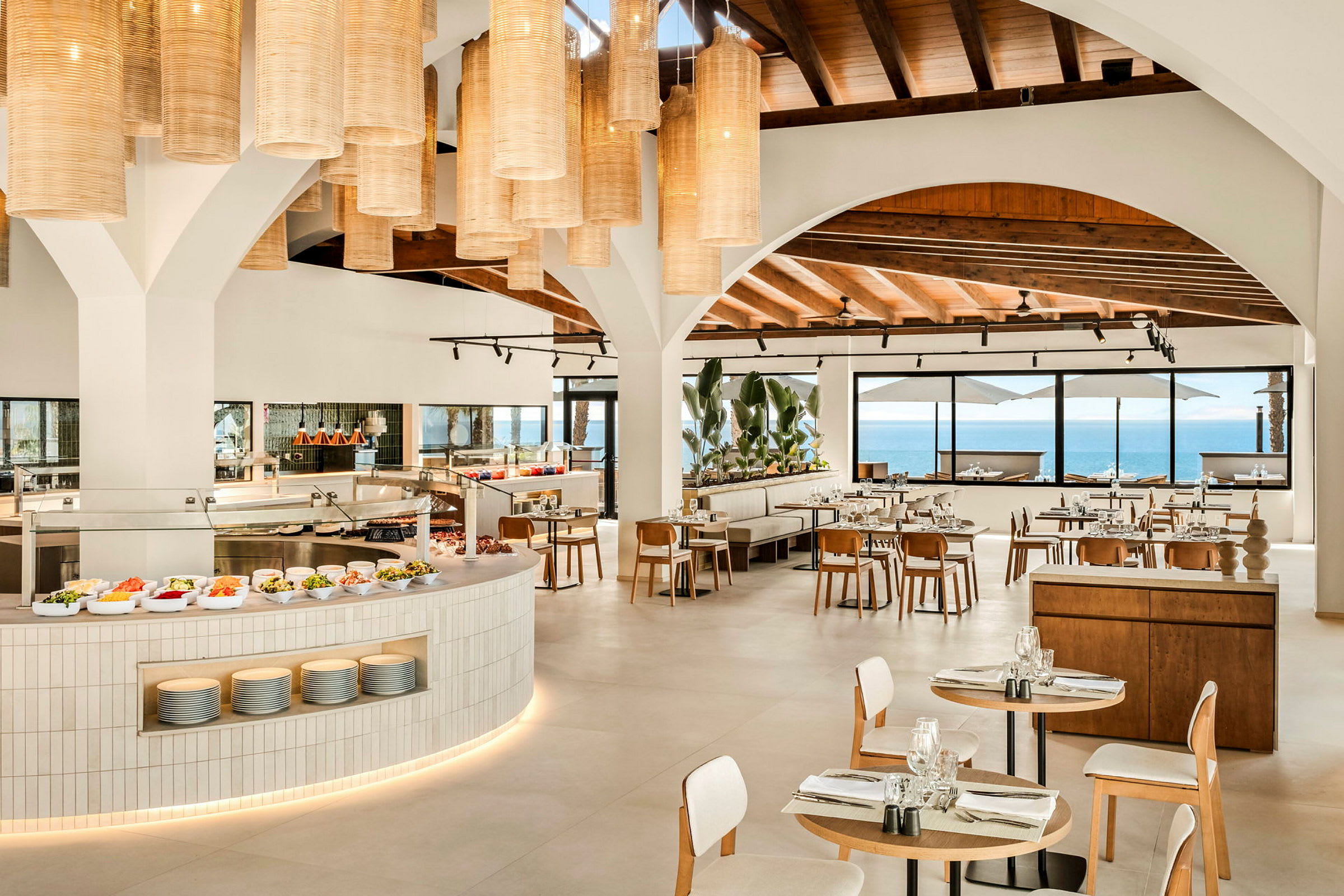 Restaurant med store buede vinduer mod havet, moderne møbler og hængende lamper i loftet. (AI-genereret tekst)