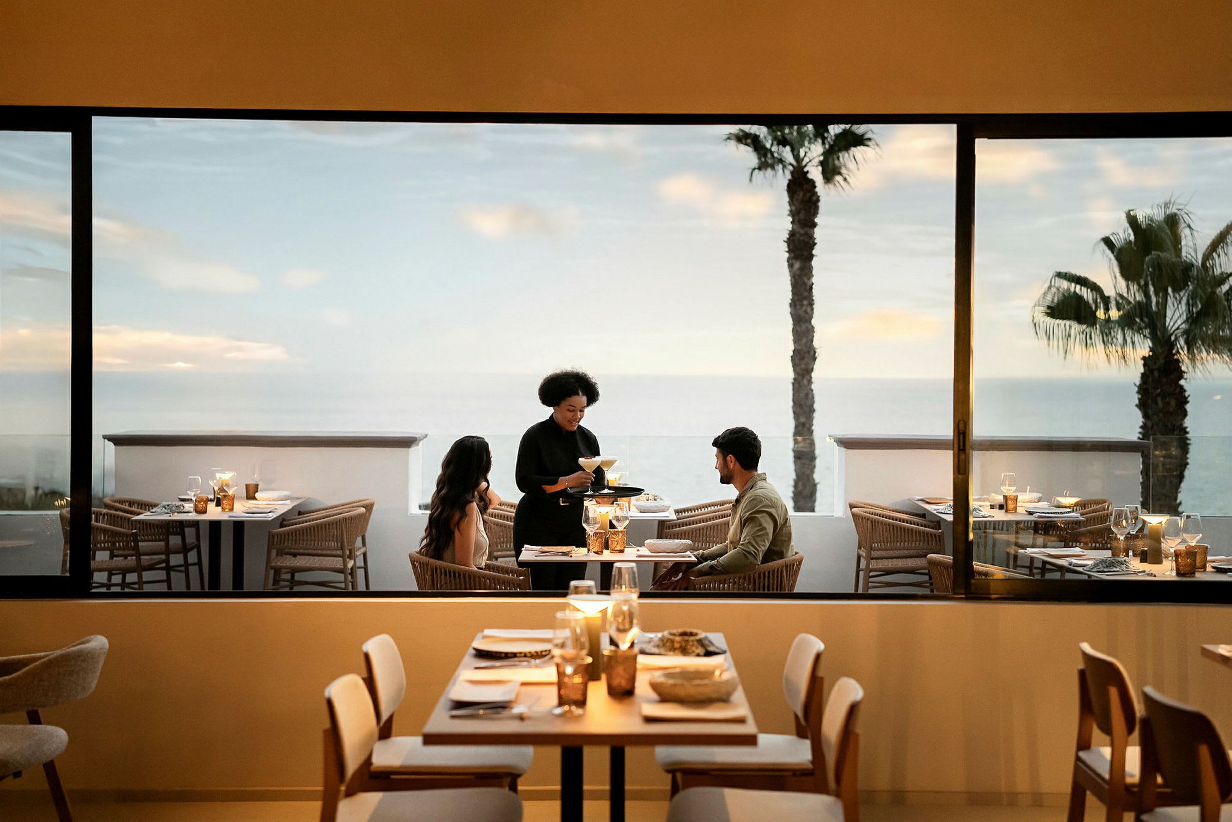En restaurant med dækkede borde, en tjener serverer et par, der sidder ved et bord. Udsigt over havet og palmer ses gennem vinduet. (AI-genereret tekst)