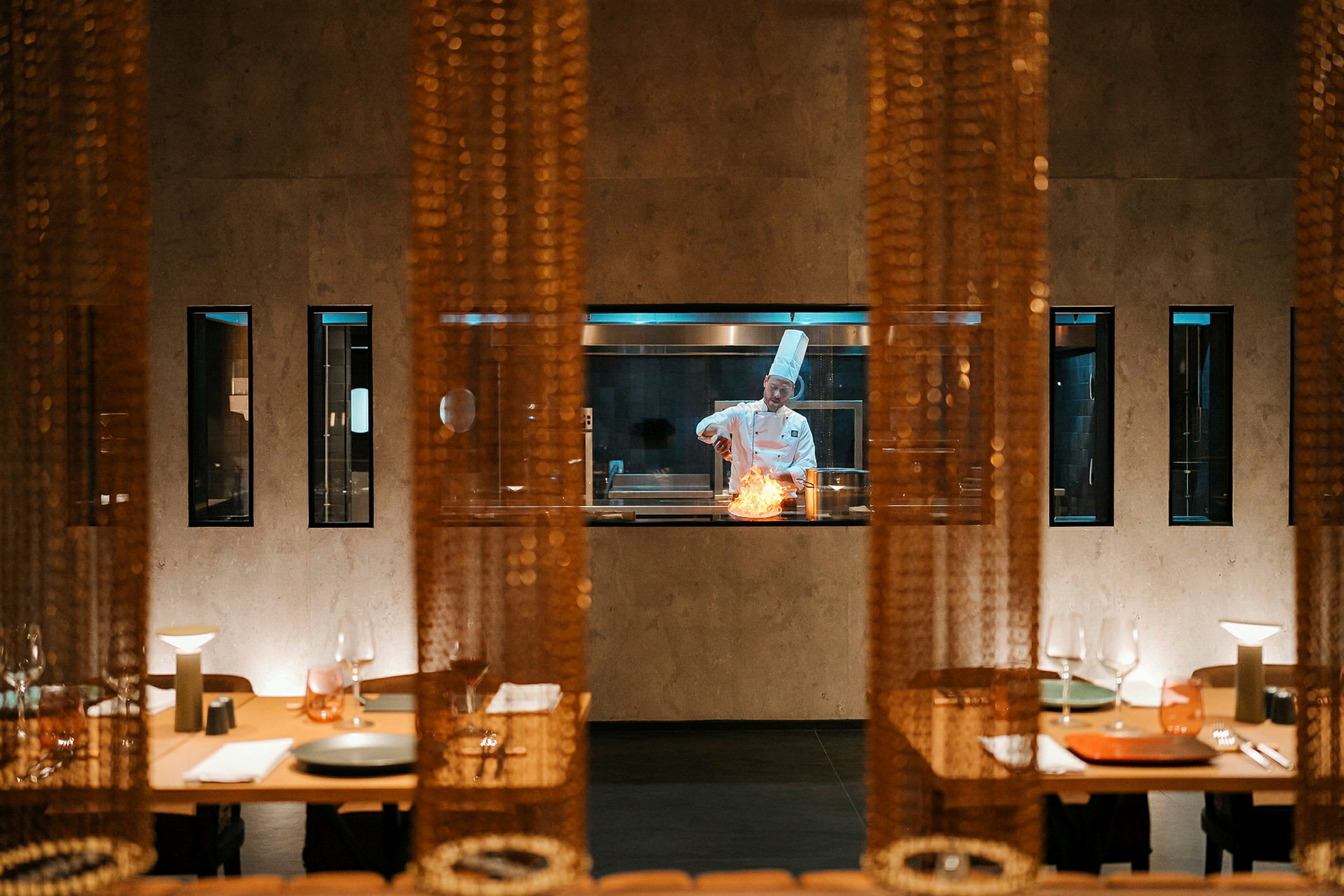 En kok ses gennem en glasvæg i en restaurantmiljø, arbejder ved en gryde med en flamme. Forgrunden har opdækkede borde. (AI-genereret tekst)