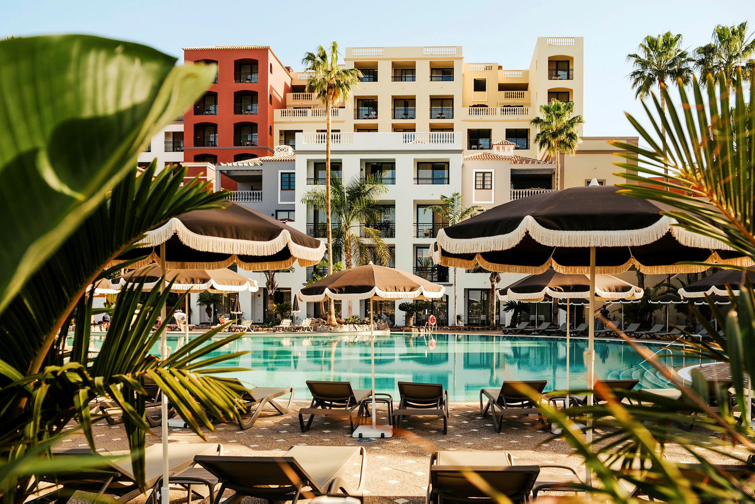 En udendørs pool foran et hotel med parasoller og liggestole omkring. (AI-genereret tekst)