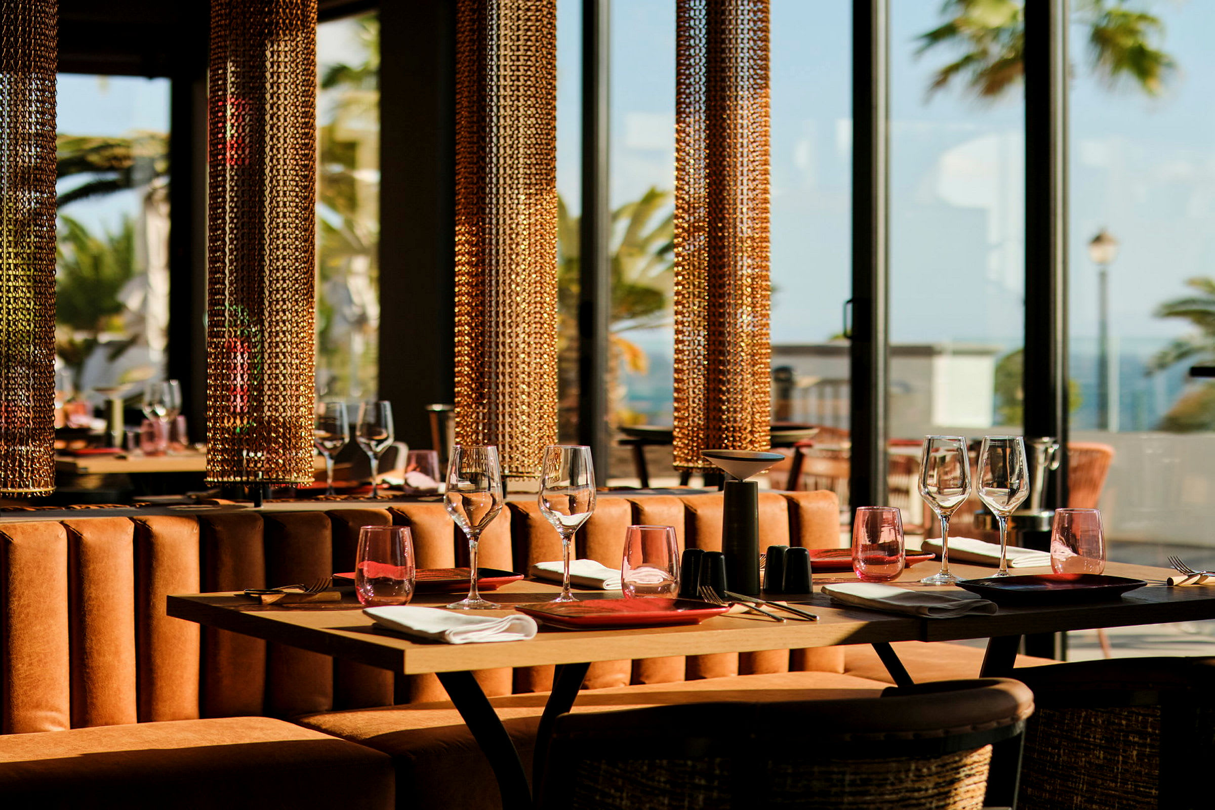 Et elegant restaurantbord dækket med glas, tallerkener og servietter, omgivet af glasvægge med udsigt over palmer. (AI-genereret tekst)