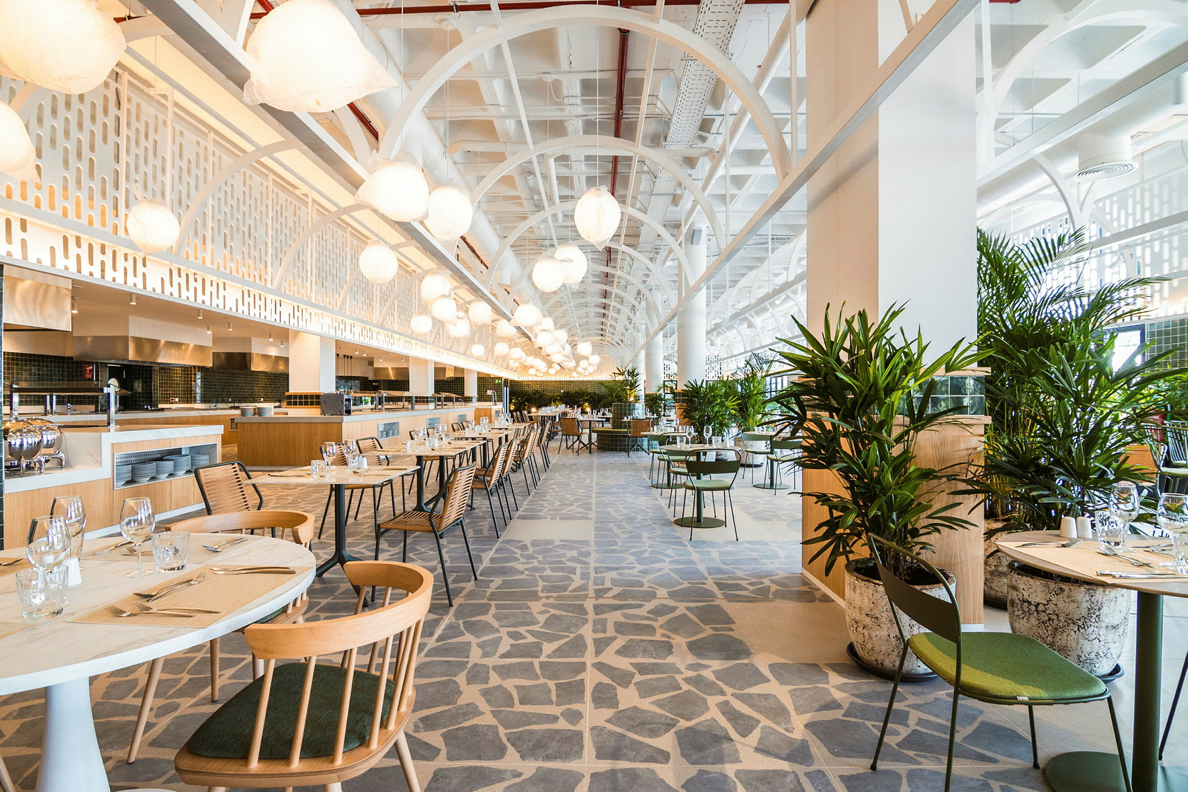 En moderne og lys restaurant med borde og stole samt grønne planter. Loftshøjde og belysning bidrager til en luftig atmosfære. (AI-genereret tekst)