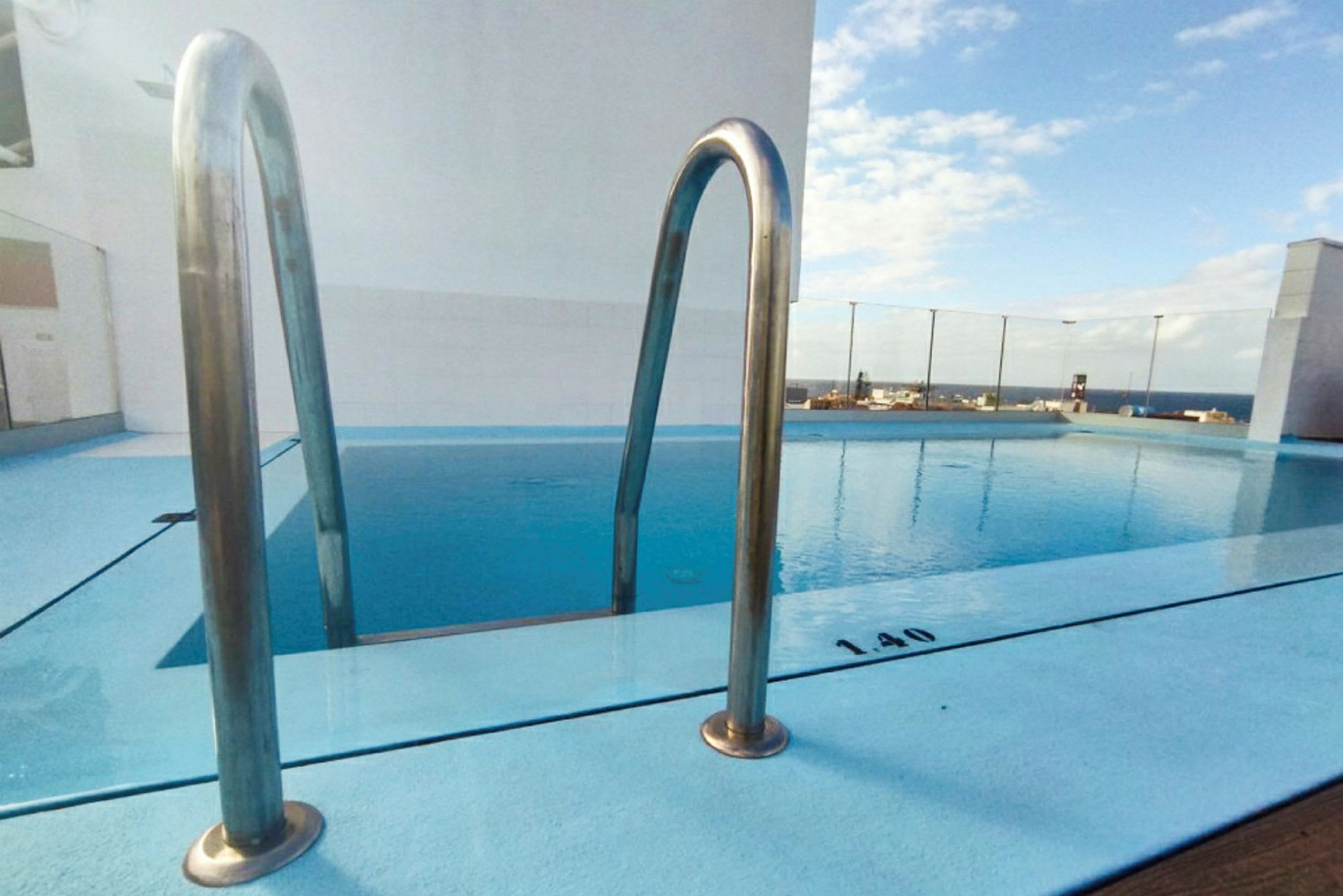 Udendørs swimmingpool med blåt vand og en rustfri poolstige, med en gennemsigtig glassvæg bagved og udsigt over landskabet. (AI-genereret tekst)