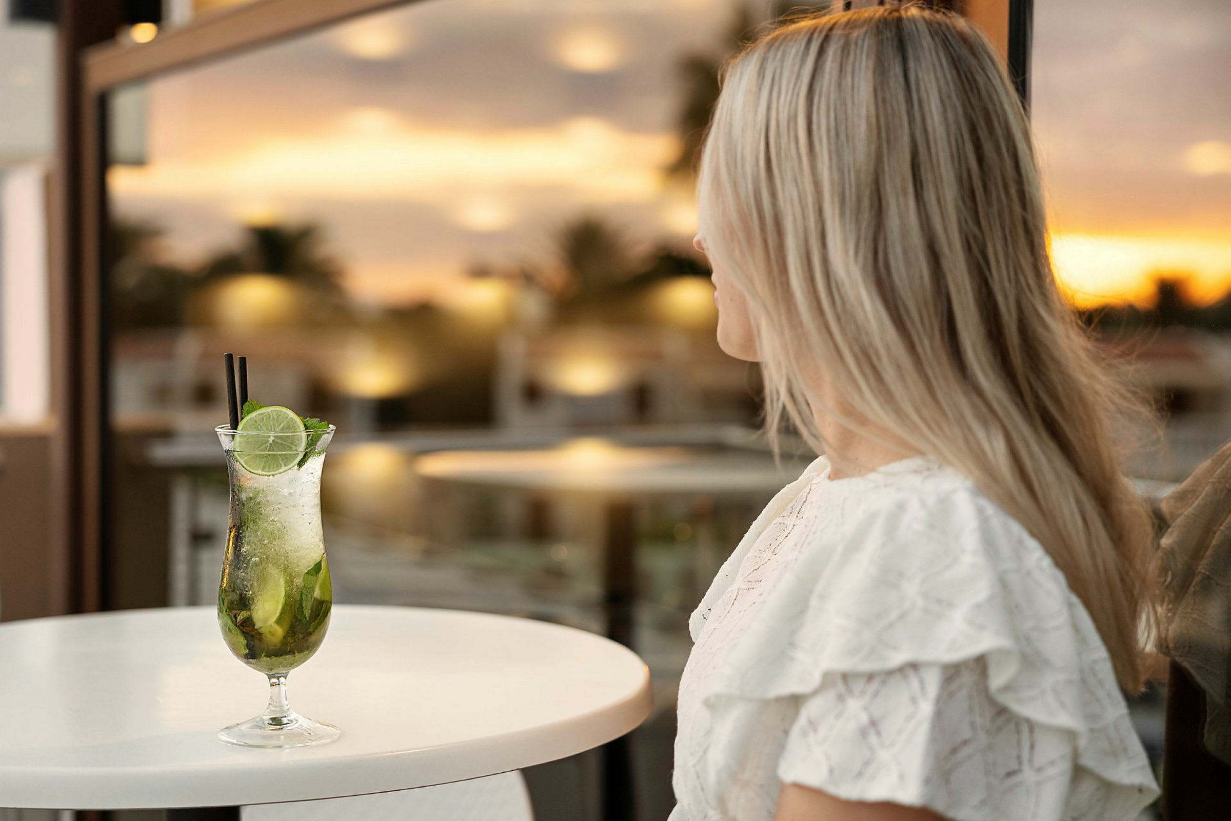 Kvinde med langt lyst hår sidder ved et bord med en cocktail foran sig, baggrunden er en solnedgang. (AI-genereret tekst)