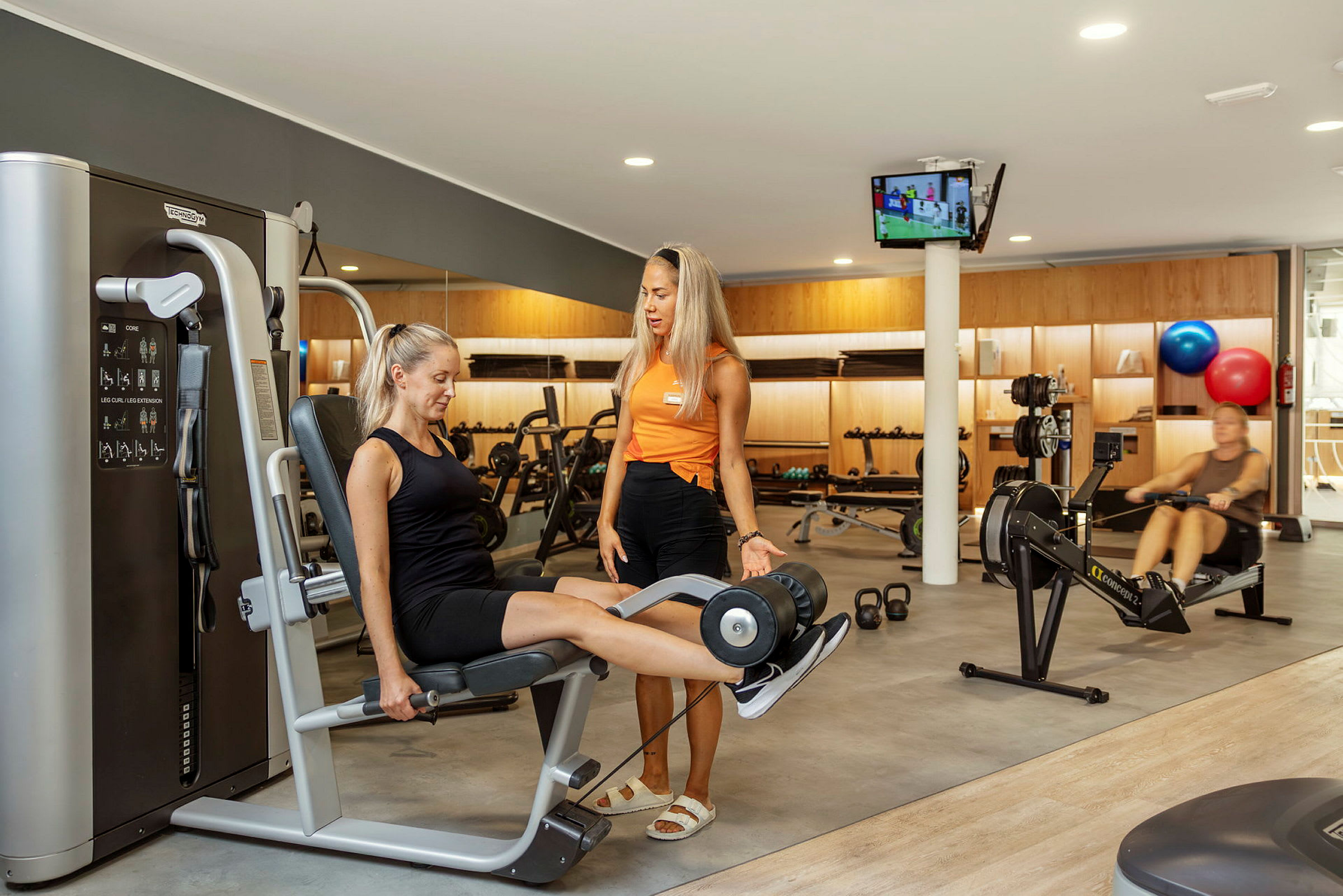 Miljøbillede fra et fitnesscenter med træningsudstyr og personer, der træner. (AI-genereret tekst)