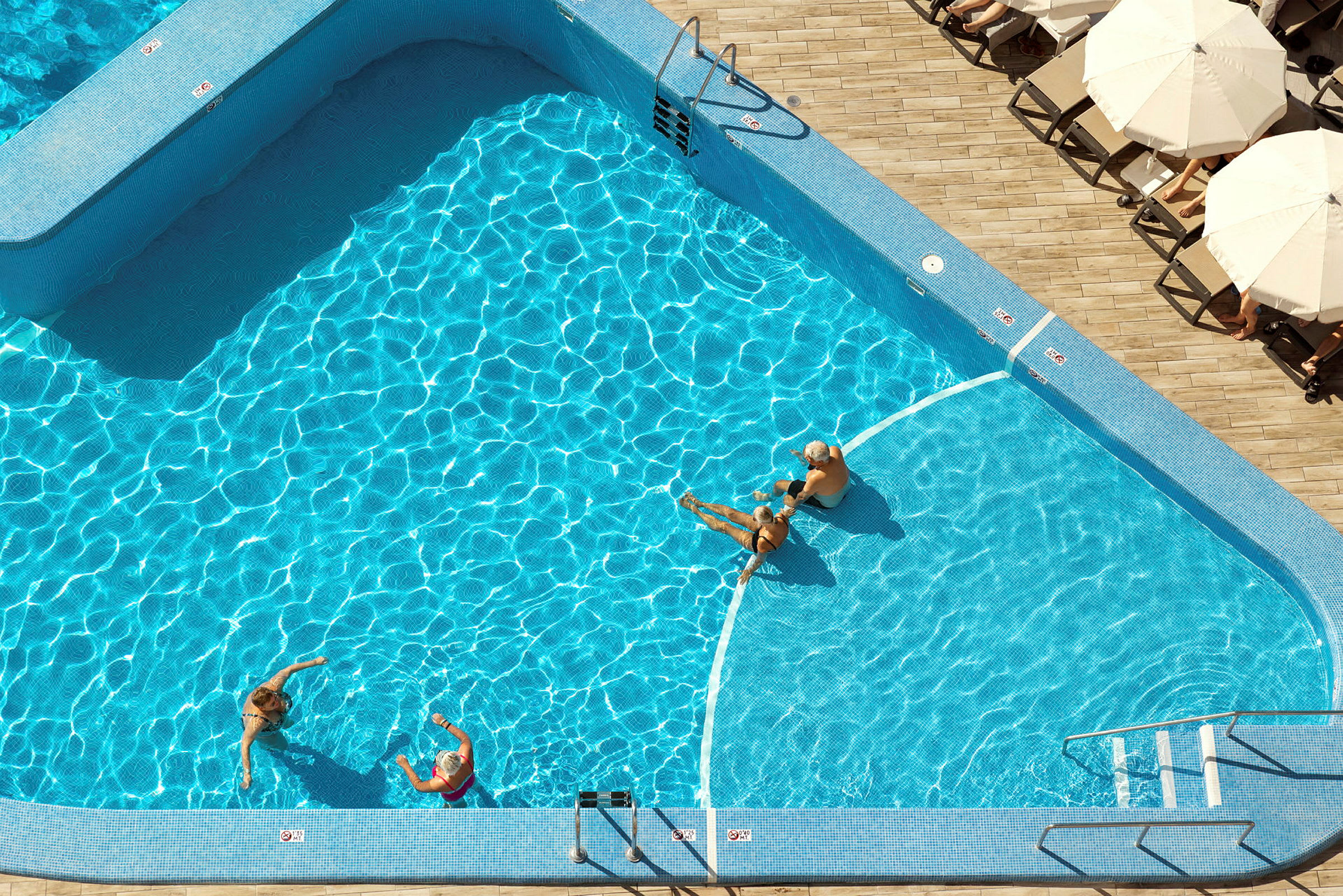 En udendørs pool med klart blåt vand, omgivet af liggestole og parasoller. Nogle mennesker bader. (AI-genereret tekst)