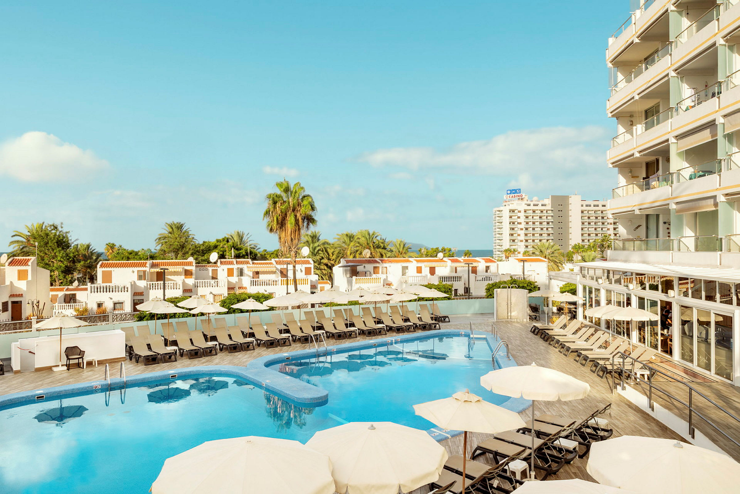 Udsigt over hotelpool med liggestole og parasoller, omgivet af bygninger og palmer. (AI-genereret tekst)